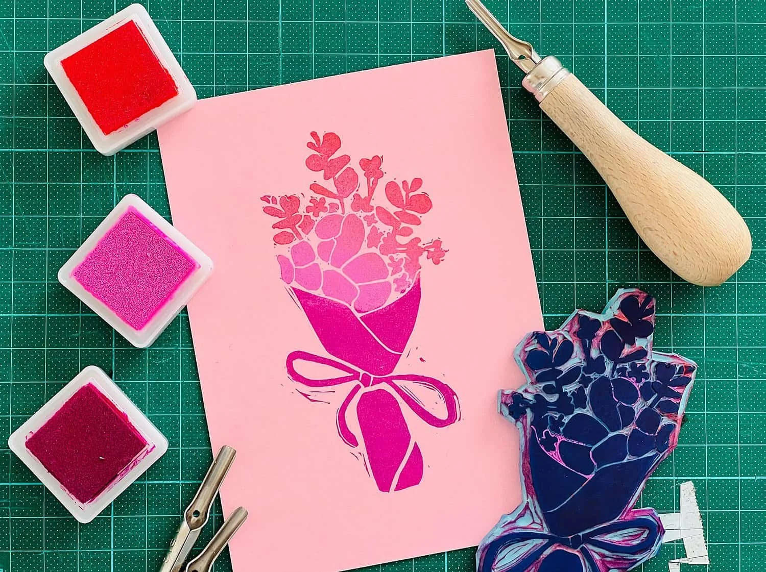 Handgesdruckte Karte eines Blumenstraußes, umgeben von Stempeln, Schneidewerkzeug, Schere und Stempelkissen auf einem grünen Schneidebrett.