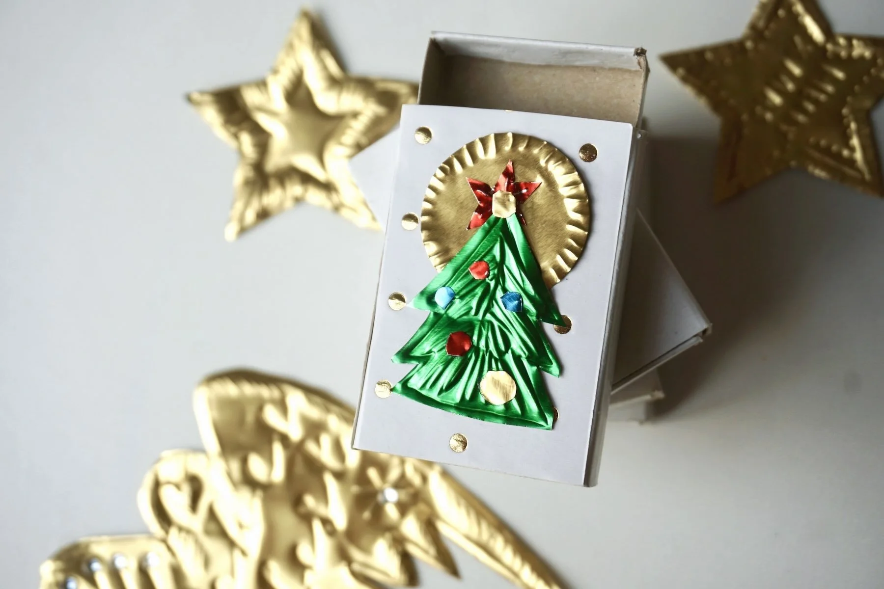 weiße Schachtel mit Tannenbaum Motiv drauf ausgeschnitten aus der Prägefolie mit buntem Baumschmuck und goldenem Kreis drum herum