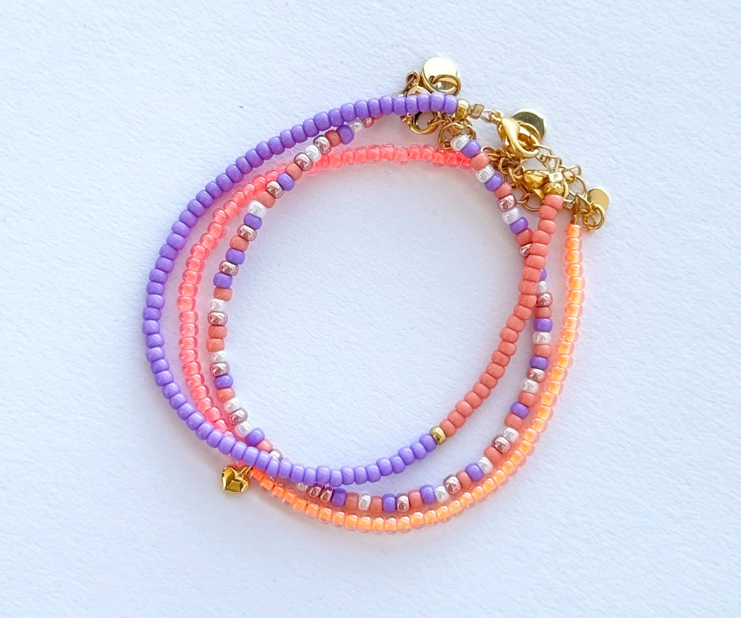 Drei bunte Musterarmbänder aus kleinen Perlen in Pastellfarben, mit goldenen Verschlüssen, auf weißem Hintergrund.