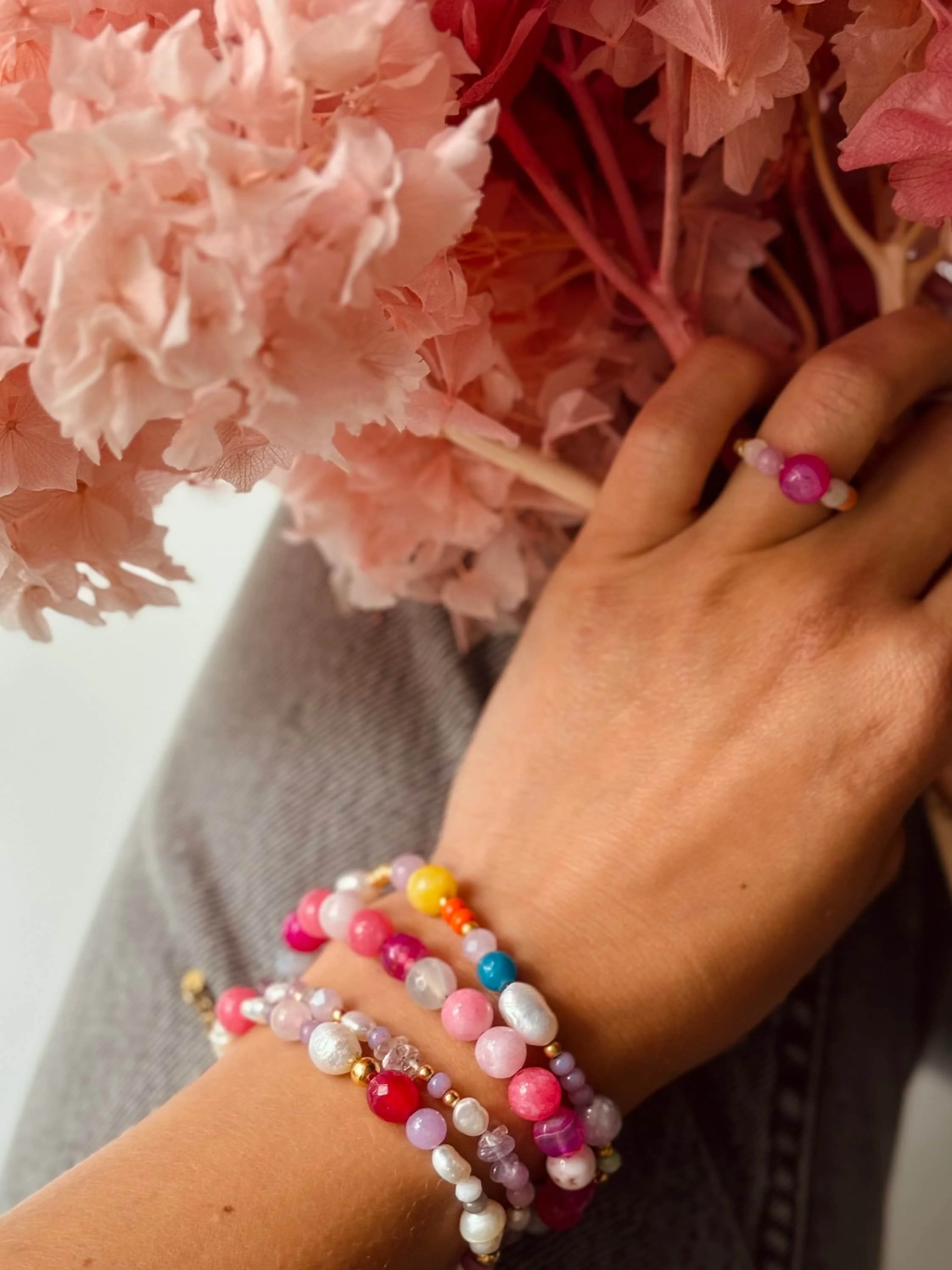 Eine Hand trägt mehrere bunte Perlenarmbänder und eine Perlenring, neben rosa Trockenblumen.