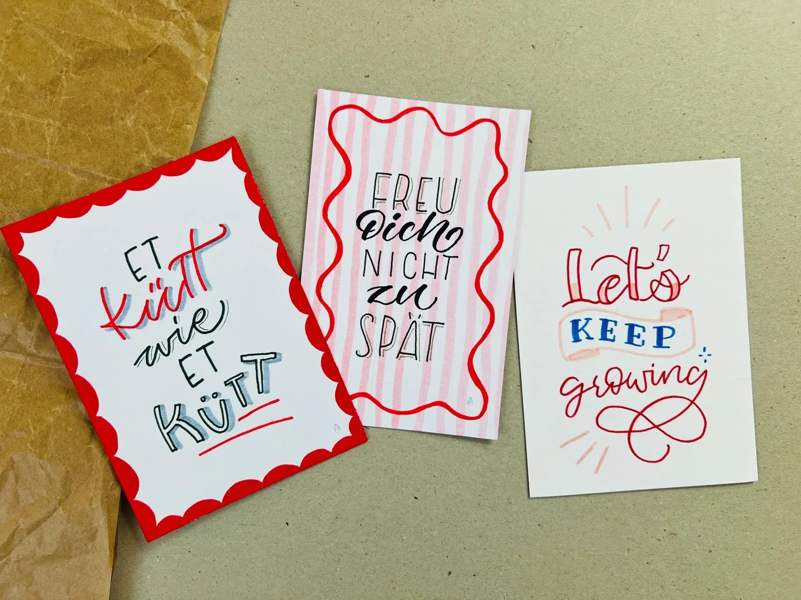 Drei handschriftliche Karten mit Sprüchen 'Et kütt wie et kütt', 'Freu dich nicht zu spät', Let's keep growing' auf weißem Papier, mit Umrandungen in rot und rosa.
