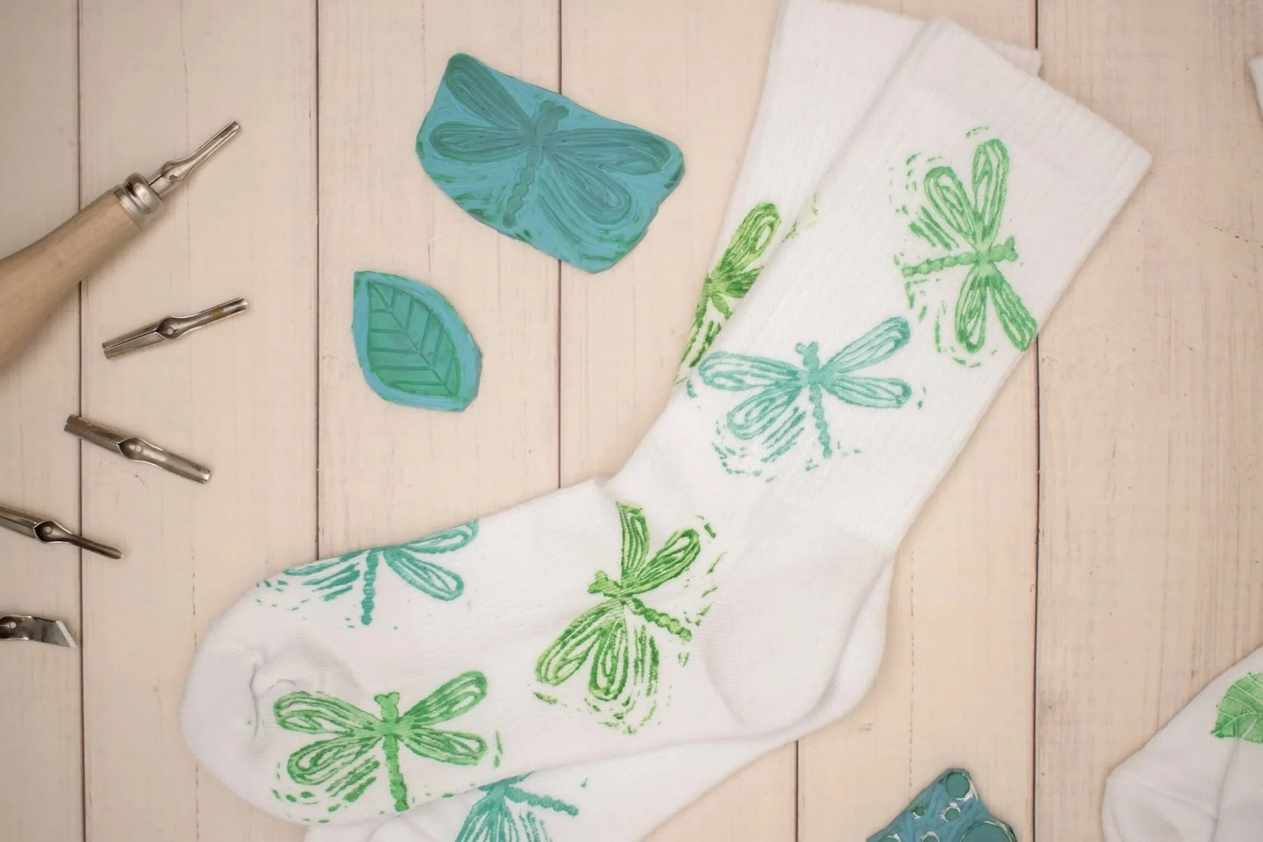 Weiße Socke mit grünen und blauen Schmetterlingsmustern, umgeben von Werkzeugen und gefärbten Schablonen mit Libellendesign auf einem Holzuntergrund.