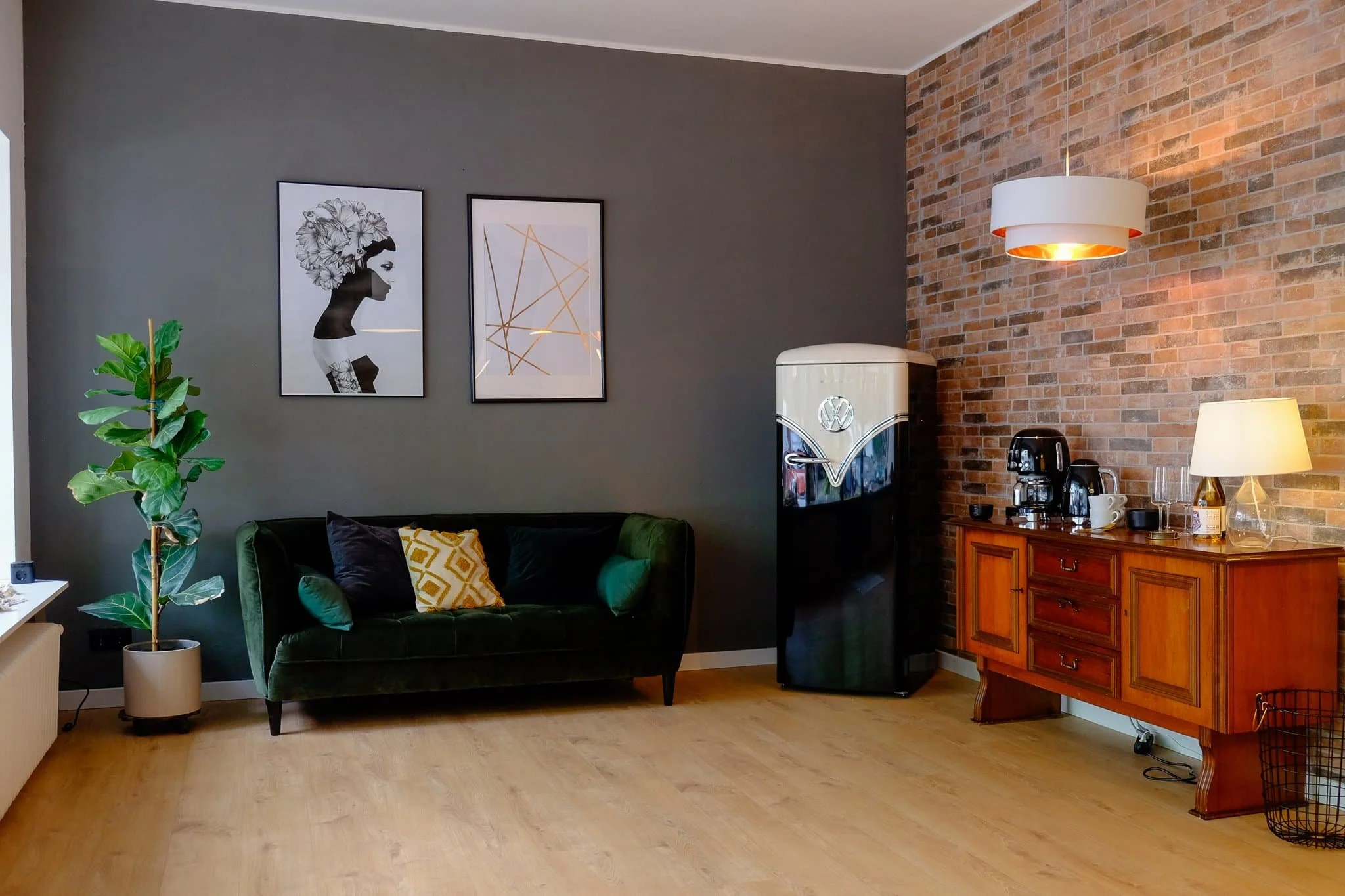 Ein modernes Zimmer mit dunkler Wand und einer Ziegelwand, grüner Samtsitzbank, Pflanzen, Wandbilder, Kühlschrank, Kaffeemaschine, Tischlampe und Holzmöbeln.