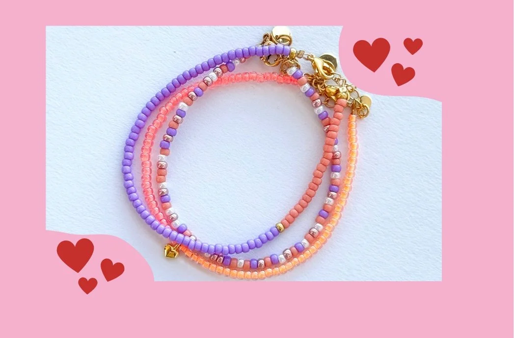 Bunte Perlenarmbänder in Rosa, Violett, Koralle mit goldfarbenen Akzenten, davor kleine Herzsymbole auf rosa Hintergrund.