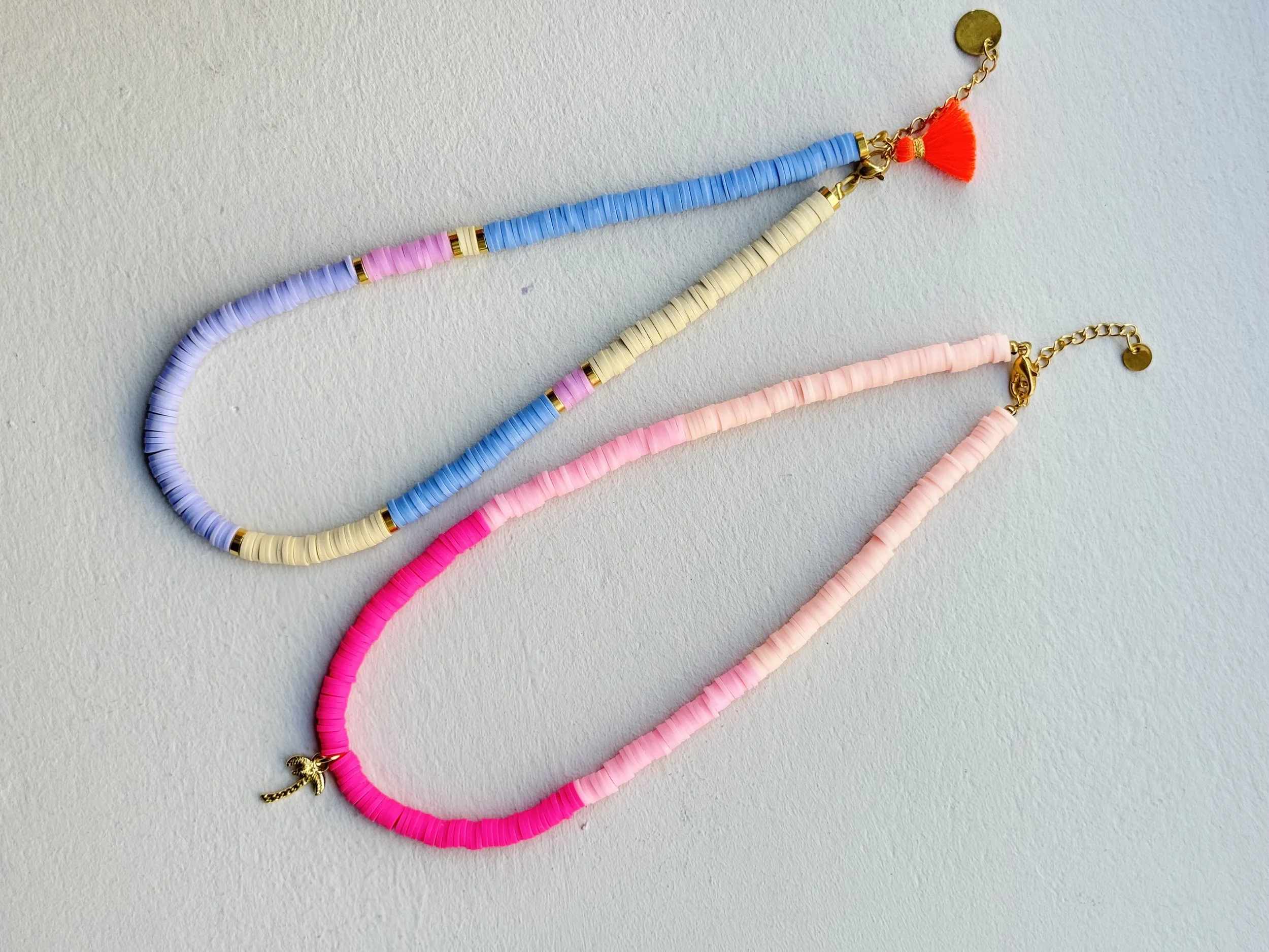 Zwei bunte Halsketten mit kleinen Scheiben, auf einem weißen Hintergrund, eine mit Pastellfarben und die andere in Neonpink, mit Goldverschluss und kleinen Anhängern