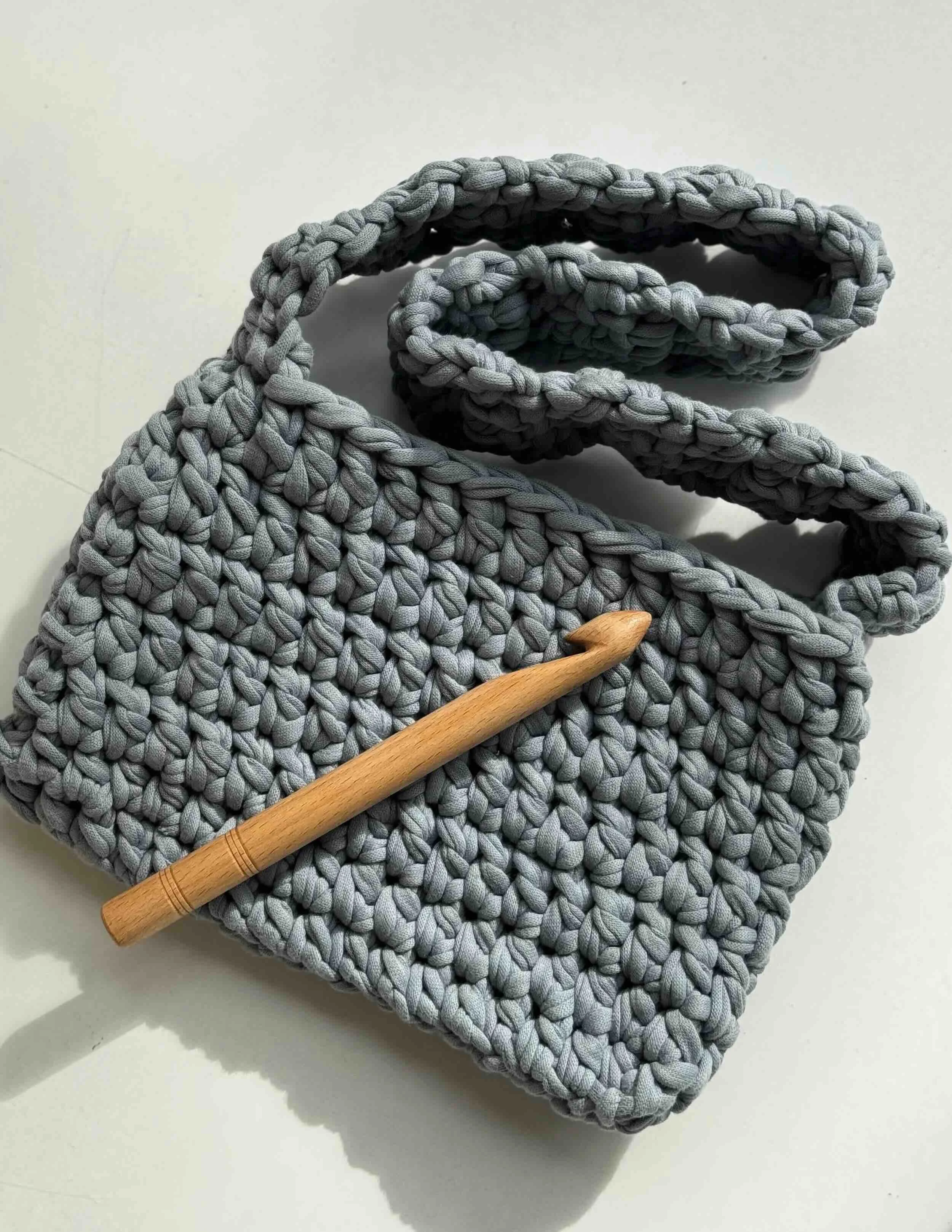 Graue gehäkelte Tasche und braune Holzhäkelnadel liegen auf einem Tisch