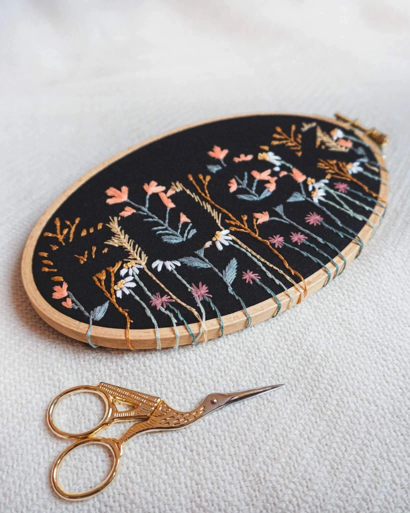 Ovale Stickarbeit auf schwarzem Stoffrahmen mit floralen Mustern in verschiedenen Farben, liegend auf einem weißen Hintergrund. Daneben liegt eine goldfarbene, scherenförmige Stickschere.