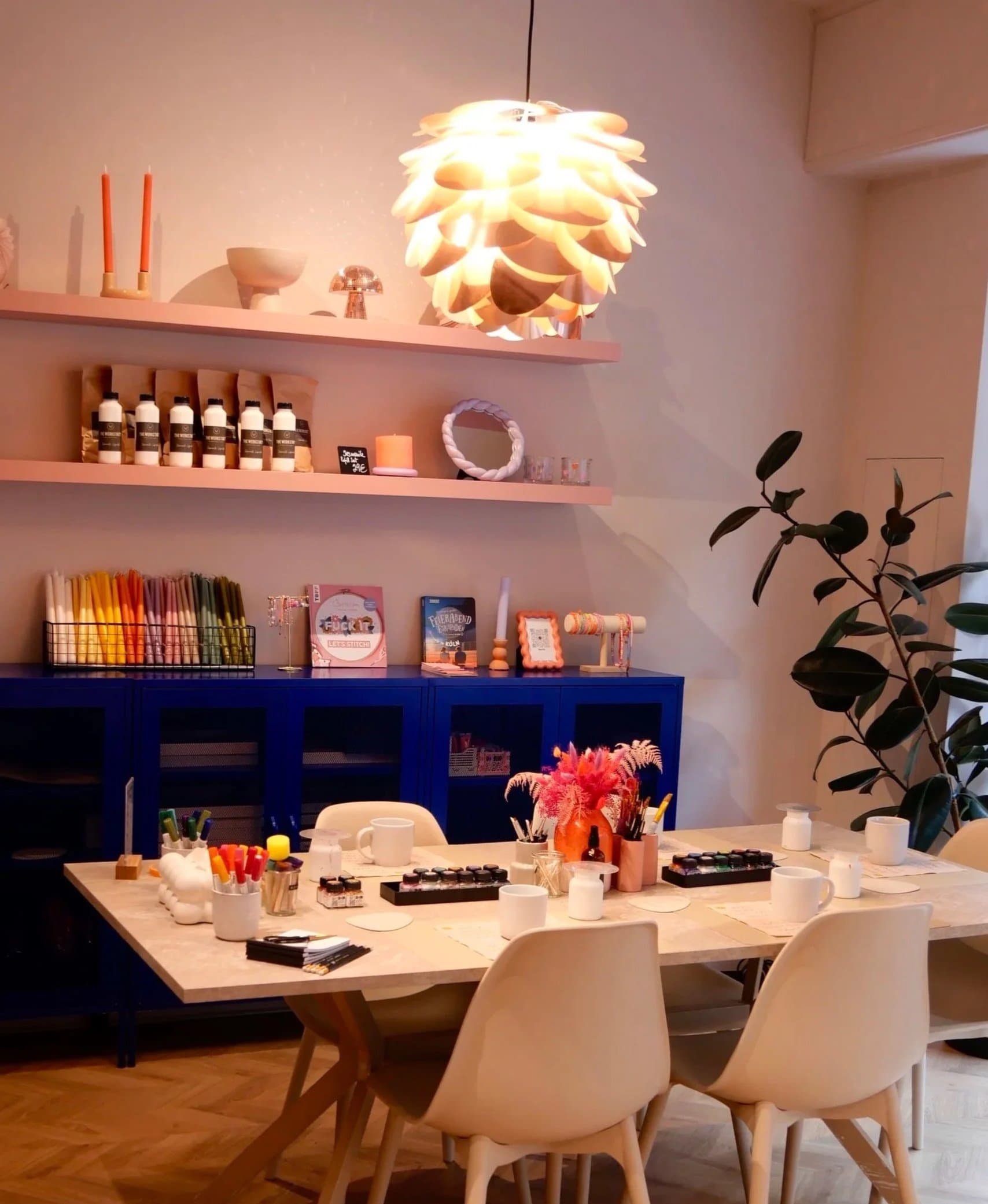 Ein modern eingerichteter Kreativraum mit einem Tisch, Stühlen, einem blauen Sideboard, Dekorationsobjekten und einem großen Deckenleuchter.