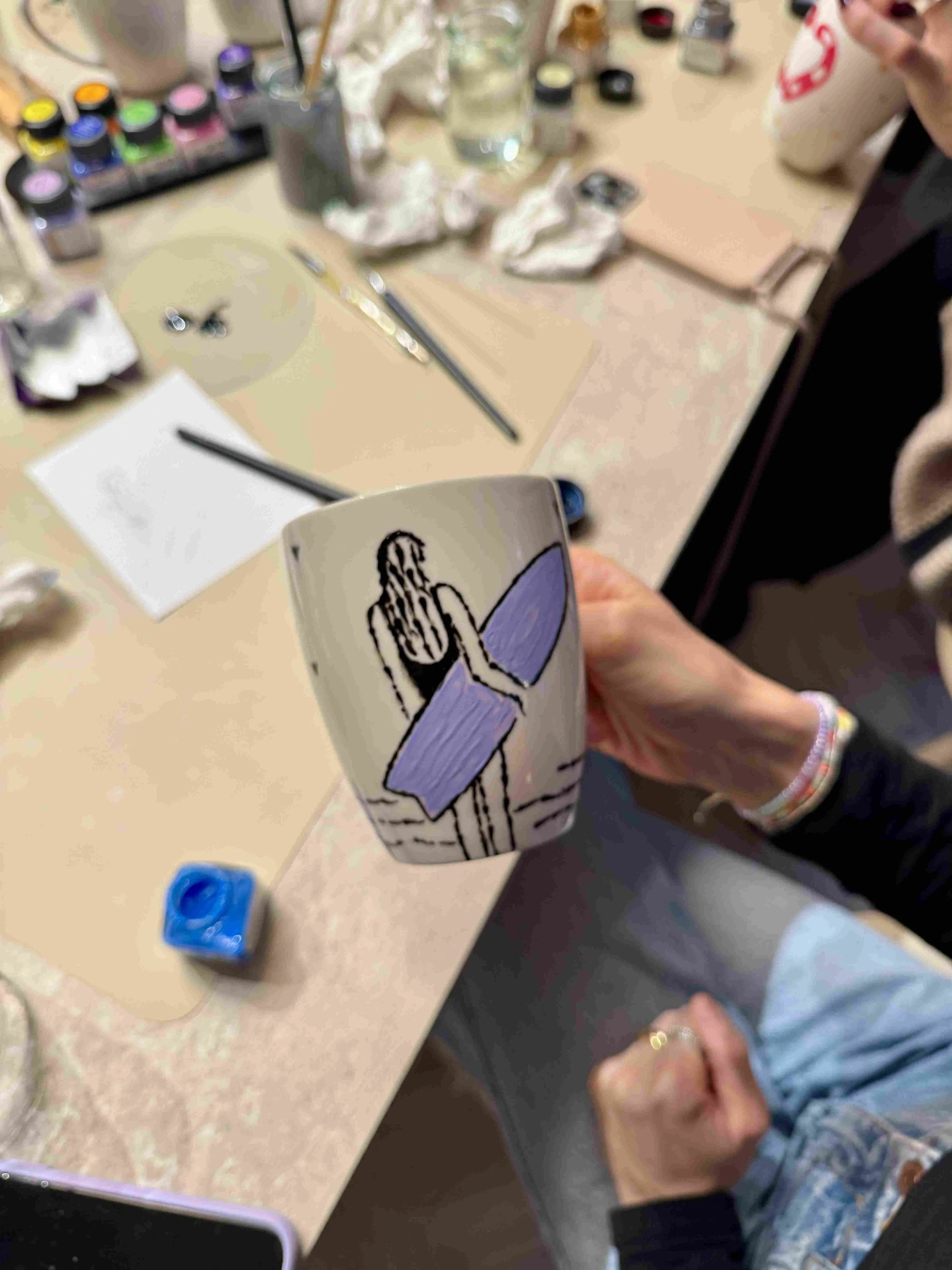 Selbst bemalte Porzellan-Tassen mit einer Illustration einer Surferin in schwarz gezeichnet mit lila Surfboard, gehalten von einer Hand über einem Tisch mit Utensilien
