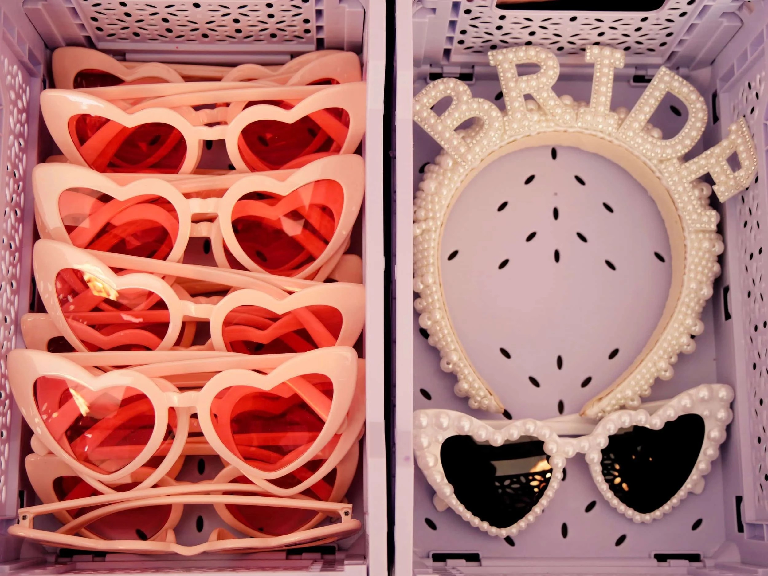 Zwei Schachteln mit rosa Sonnenbrillen in Herzform auf der linken Seite und einer DIY-Haarreif mit Perlen und dem Wort "BRIDE" sowie eine zusätzliche Herz-Sonnenbrille mit Perlenrahmen auf der rechten Seite.