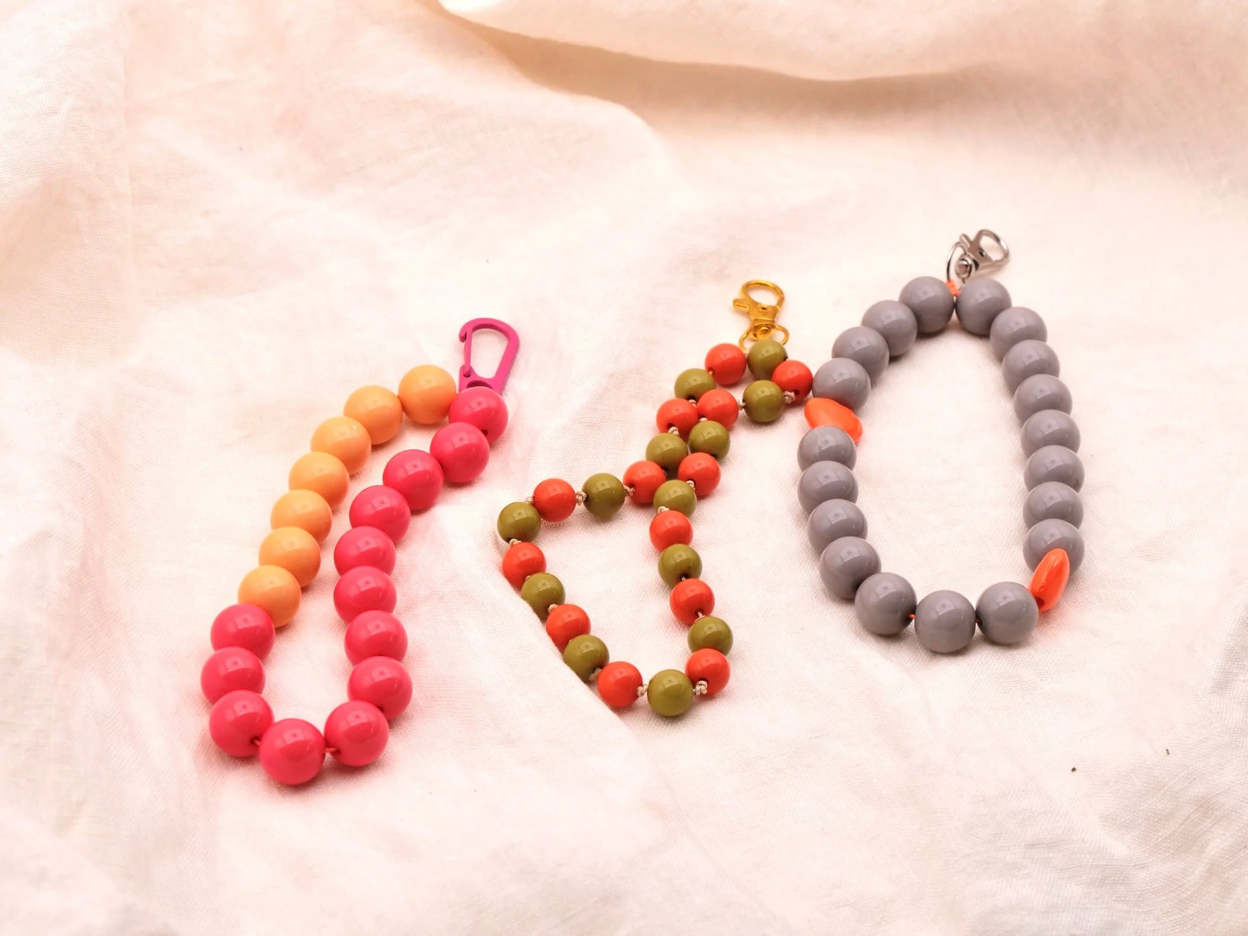 Drei bunte Schlüsselanhänger aus großen Perlen in Pink, grün und Grau auf weißem Stoffhintergrund.