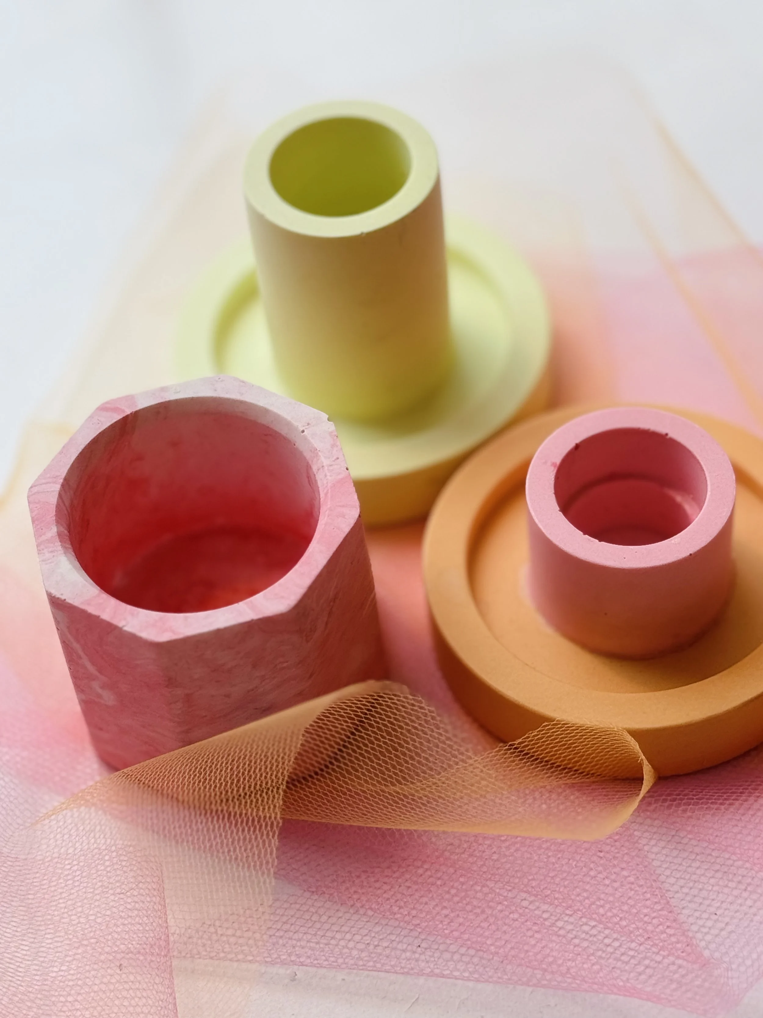 Bunte, runde und zylindrische Kerzenformen aus Jesmonite in rosa, grün und orange, auf einem rosa Tüllstoff