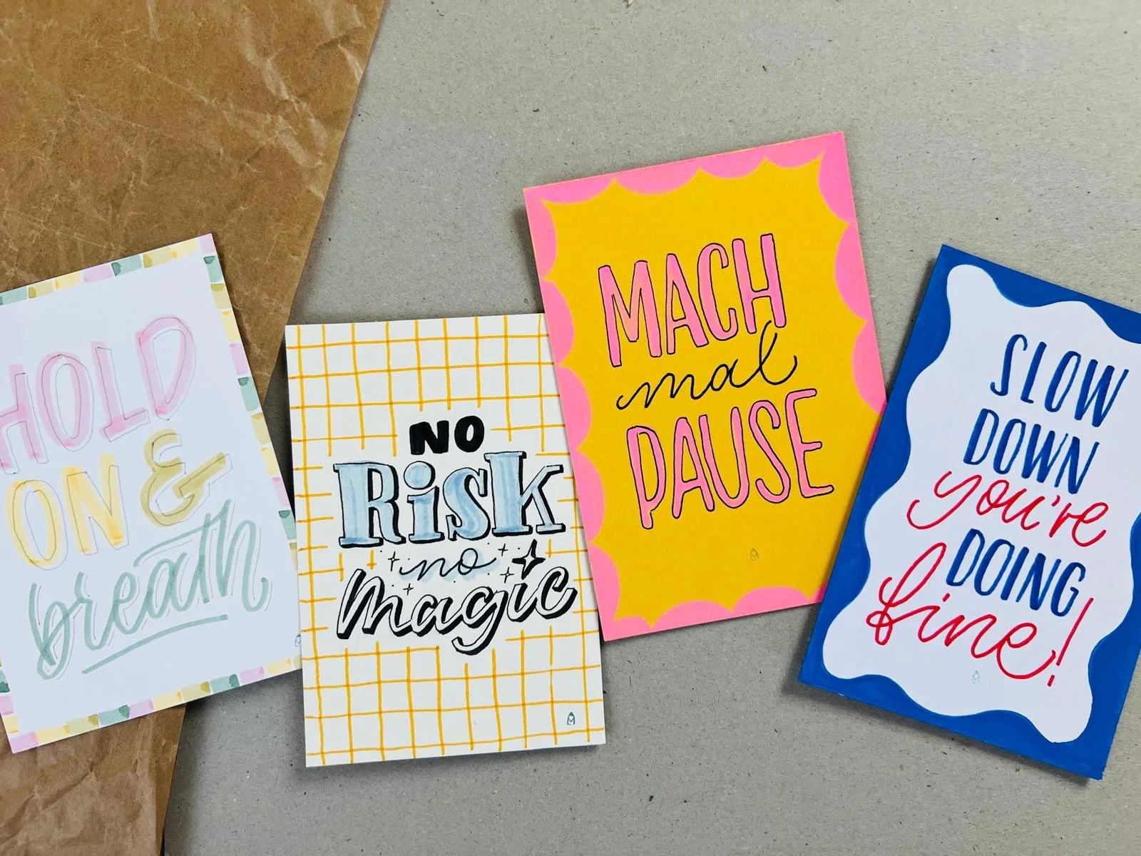 Vier handschriftliche Karten mit Sprüchen 'Hold on & breath', 'No risk no magic', Mach mal Pause', Slow down you're doing fine!' auf buntem Papier, mit Umrandungen in blau und rosa, karierten Hintergrund