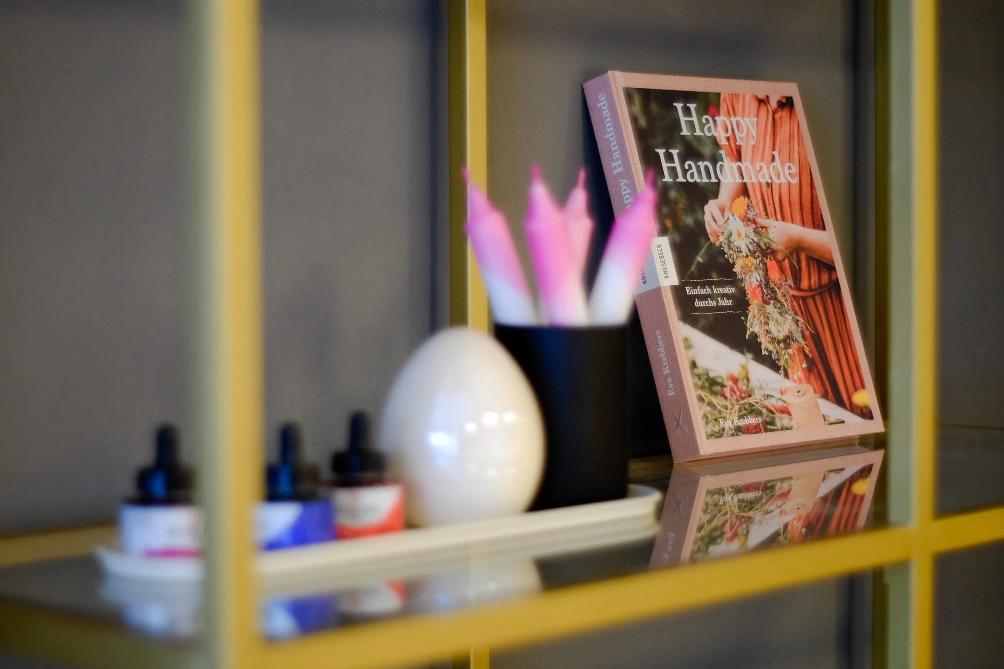 Ein Bücherregal mit einem Buch mit dem Titel 'Happy Handmade' und einer schwarzen Vase mit pinken Kerzen, daneben eine weiße Ei-Form und kleine Fläschchen mit Tinkturen.