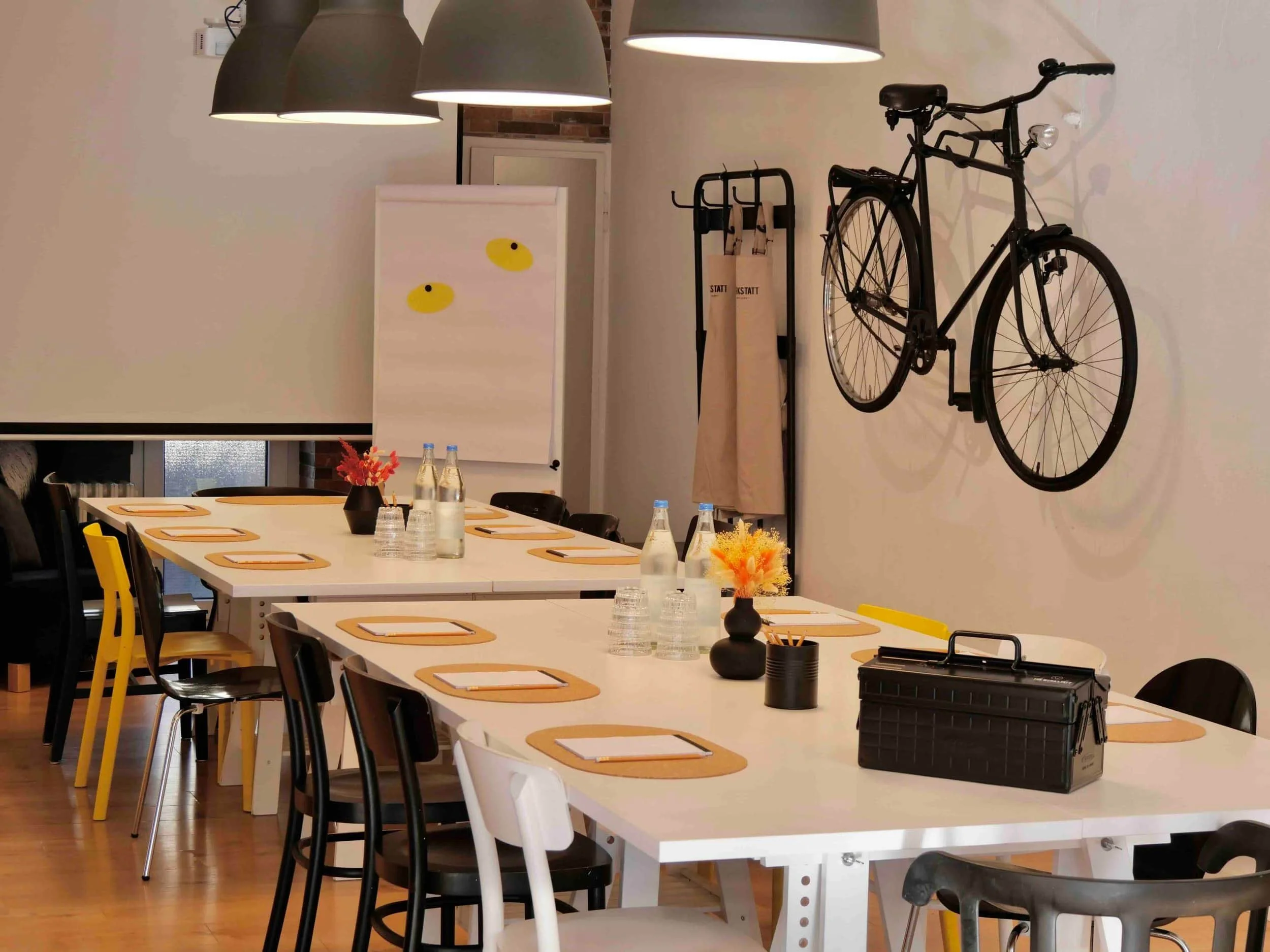 Moderner Seminarraum mit Tischtafel mit Notizblöcken, Wasserflaschen und kleinen Blumenvasen drauf, im Hintergrund sind  Leinwand, Flipchart und ein schwarzes Fahrrad