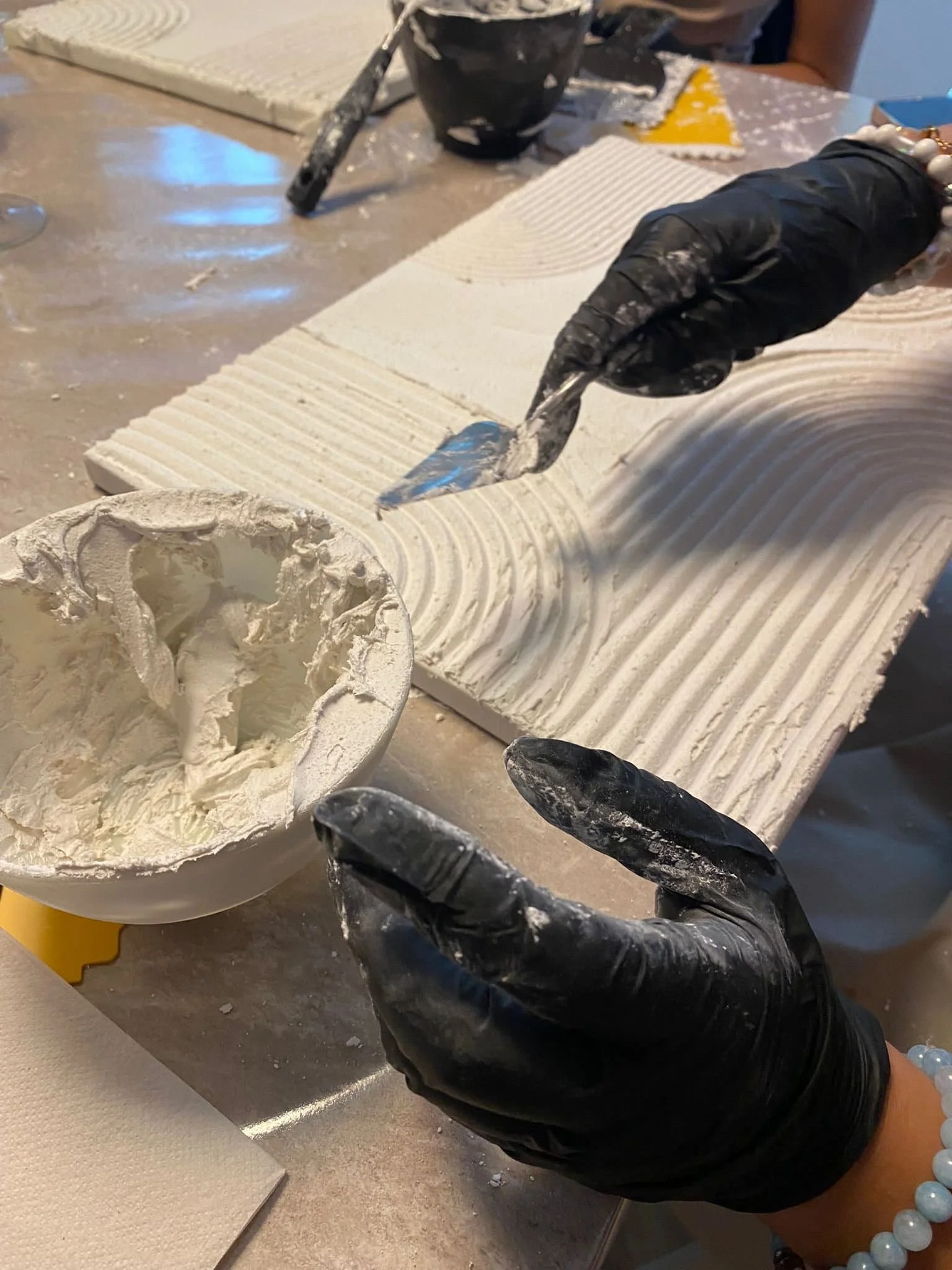 Person formt weiße Strukturpaste mit einer Spachtel. Die Person trägt schwarze Handschuhe und eine Perlenarmband.