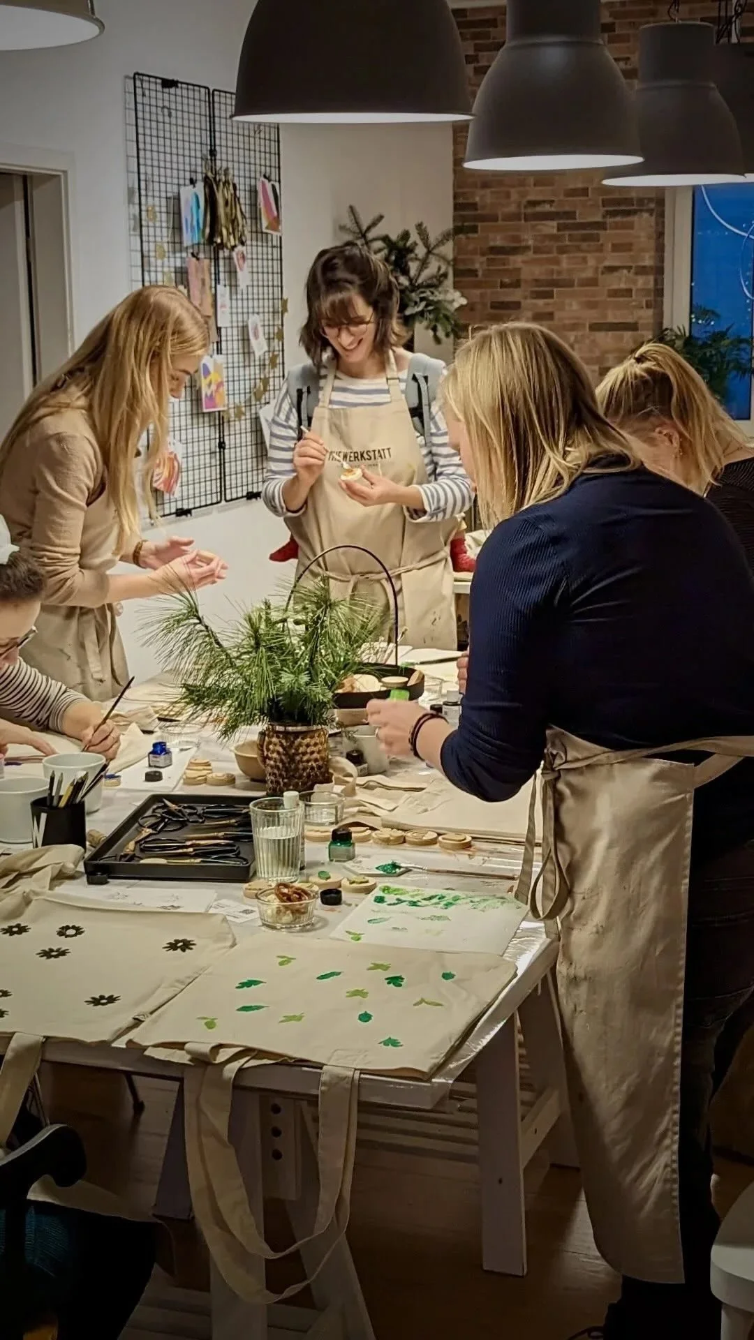 Mehrere Frauen die stehend an einem Tisch weihnachtliche Stempeln basteln, auf dem Tisch steht eine Vase mit Kieferästen sowie Pinsel, Farben, Gläser, Scheren