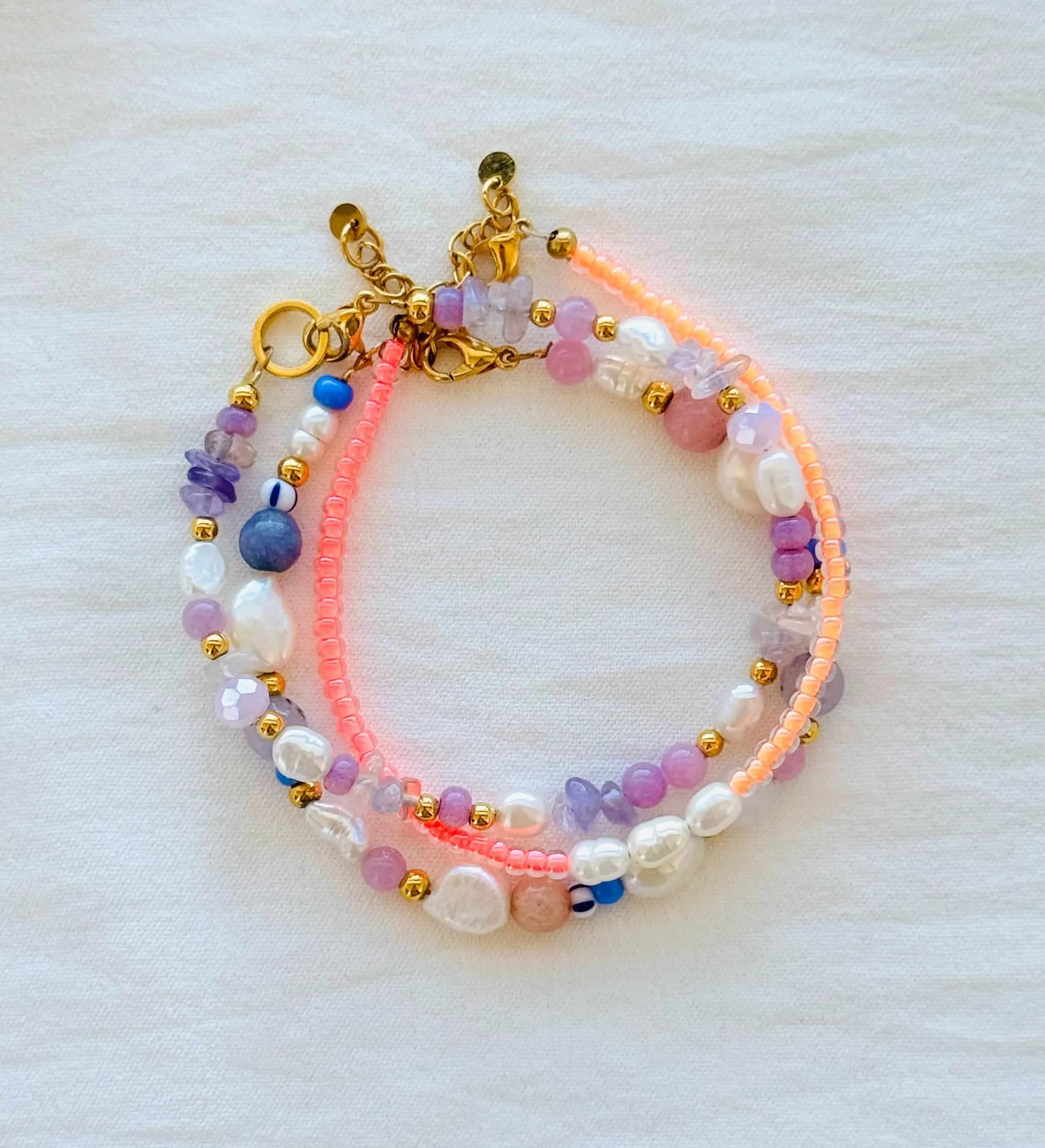 Drei Armbänder aus Miyukiperlen, Edelsteinen und Süßwasserperlen mit goldenen Verschlüssen, auf weißem Stoffuntergrund