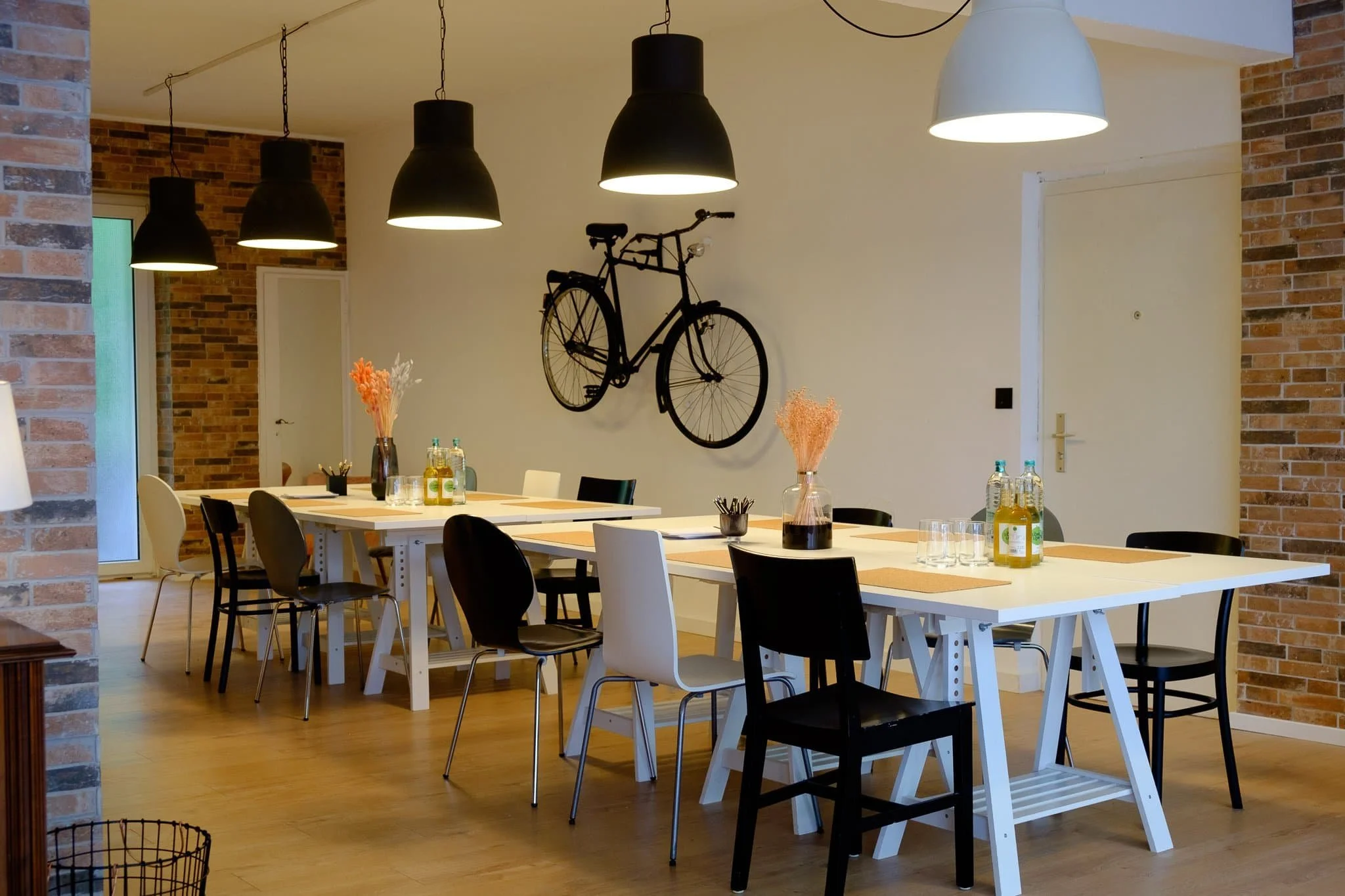 Ein modern gestalteter Raum mit weißen Tischen, schwarzen und weißen Stühlen, dekorativen Blumenvasen, Flaschen mit Getränken, schwarzen und weißen Lampen an der Decke und einer schwarzen Fahrraddekorfigur an einer weißen Wand.