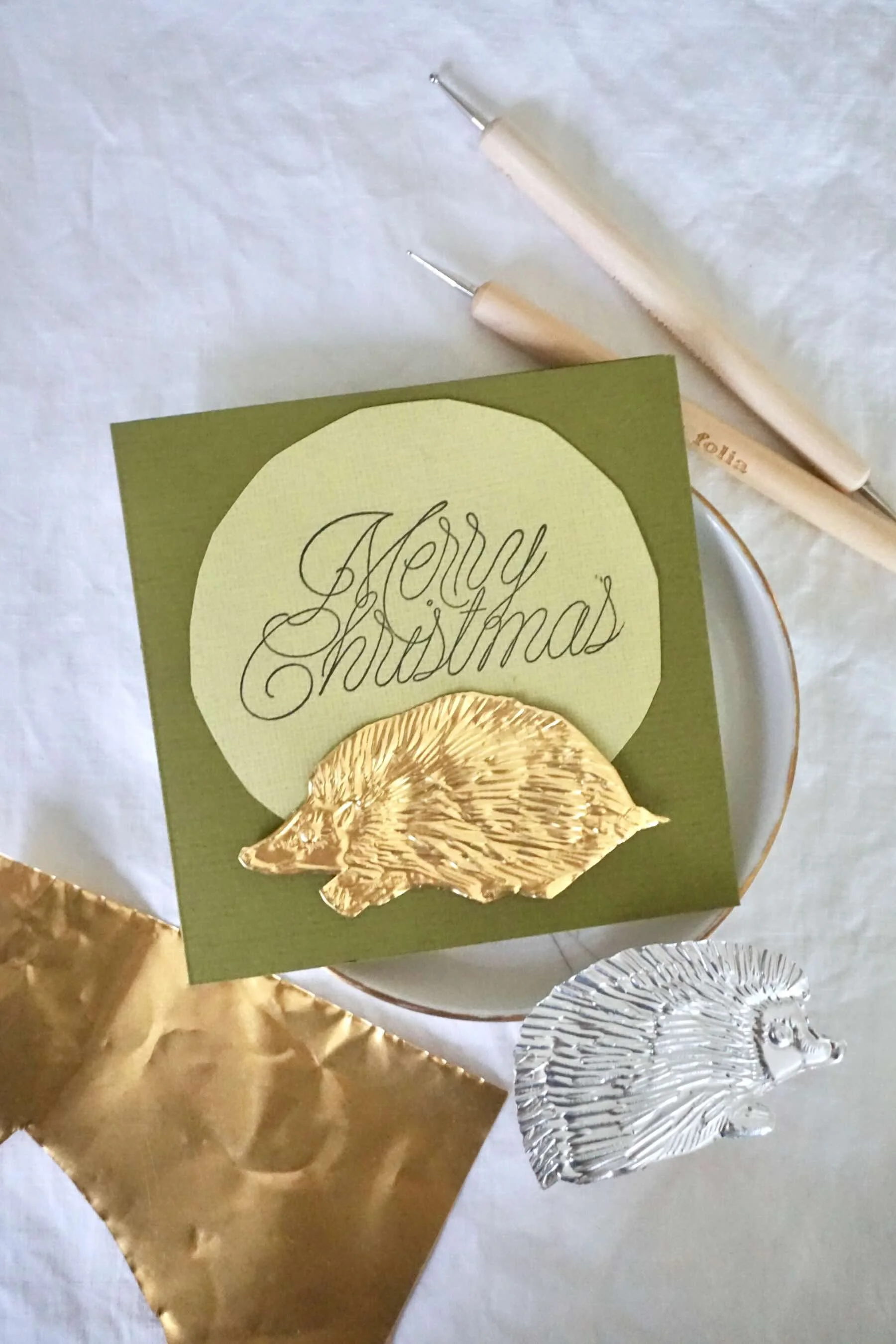 Olivgrüne Karte mit Spruch 'Merry Christmas' und einem Igel aus goldener Prägefolie