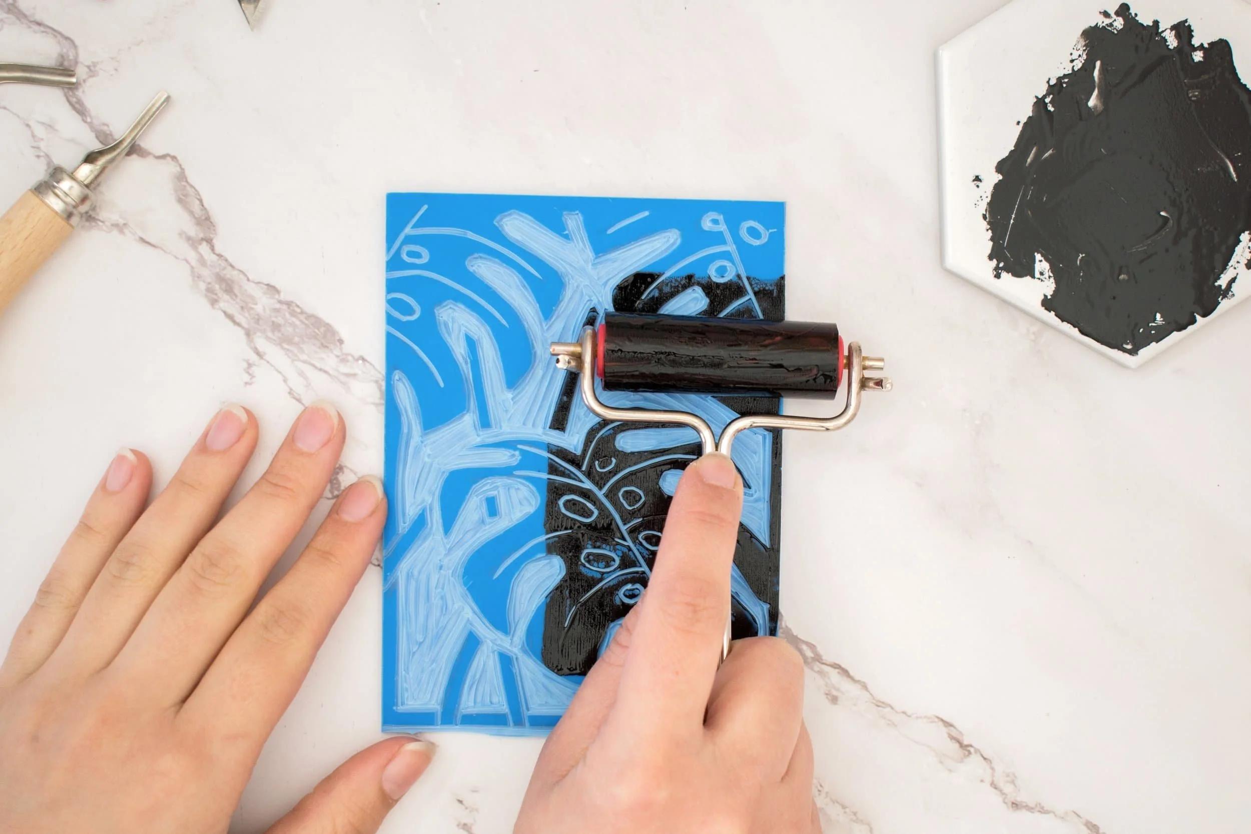 Linolschnitt von Monstera Blättern, die mit einer Walze mit schwarzer Farbe bestrichen werden auf schwarz-weiß marmoriertem Untergrund