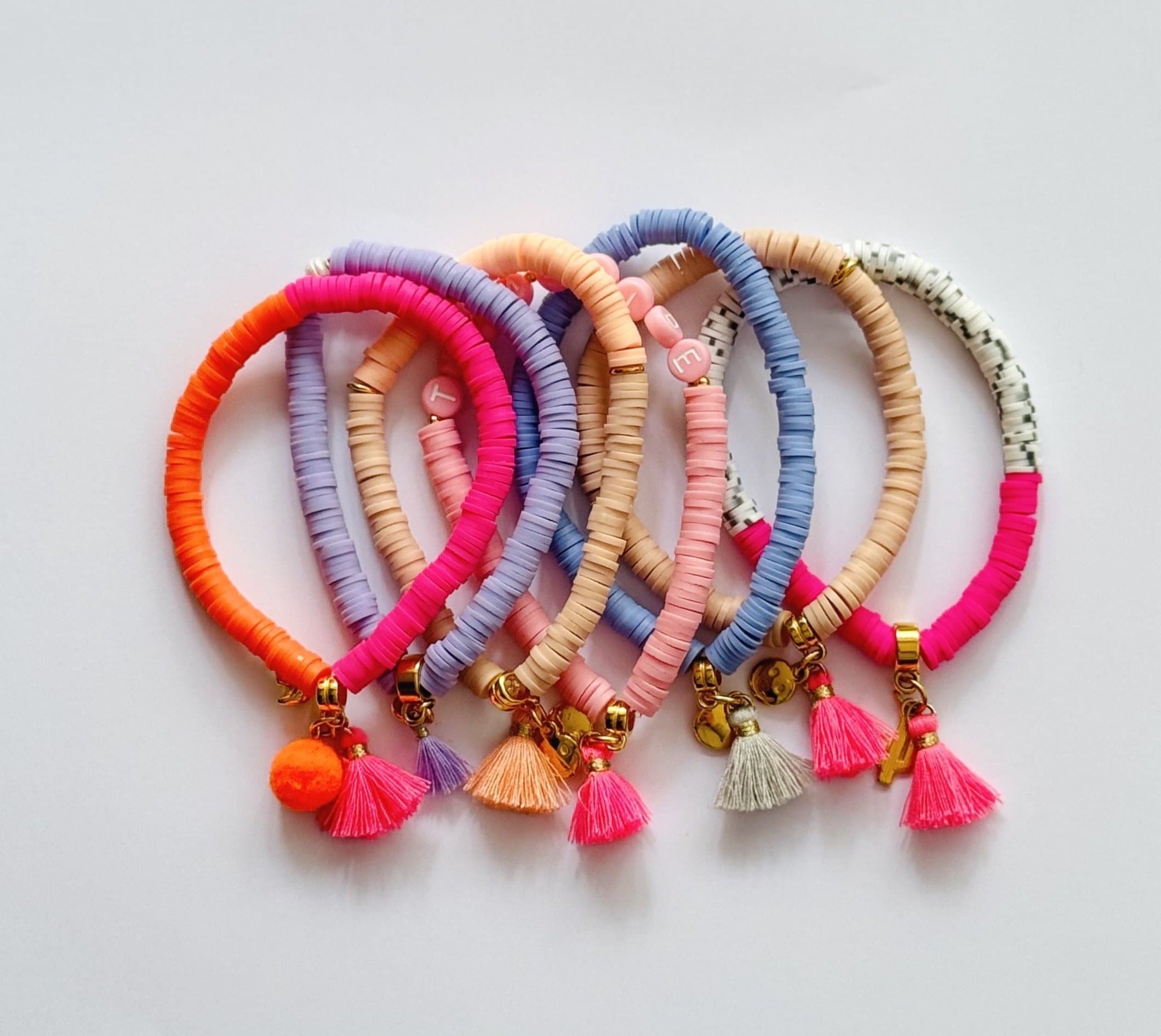 Sechs bunte Armbänder mit Quaste und Buchstaben-Anhängern auf weißem Hintergrund.