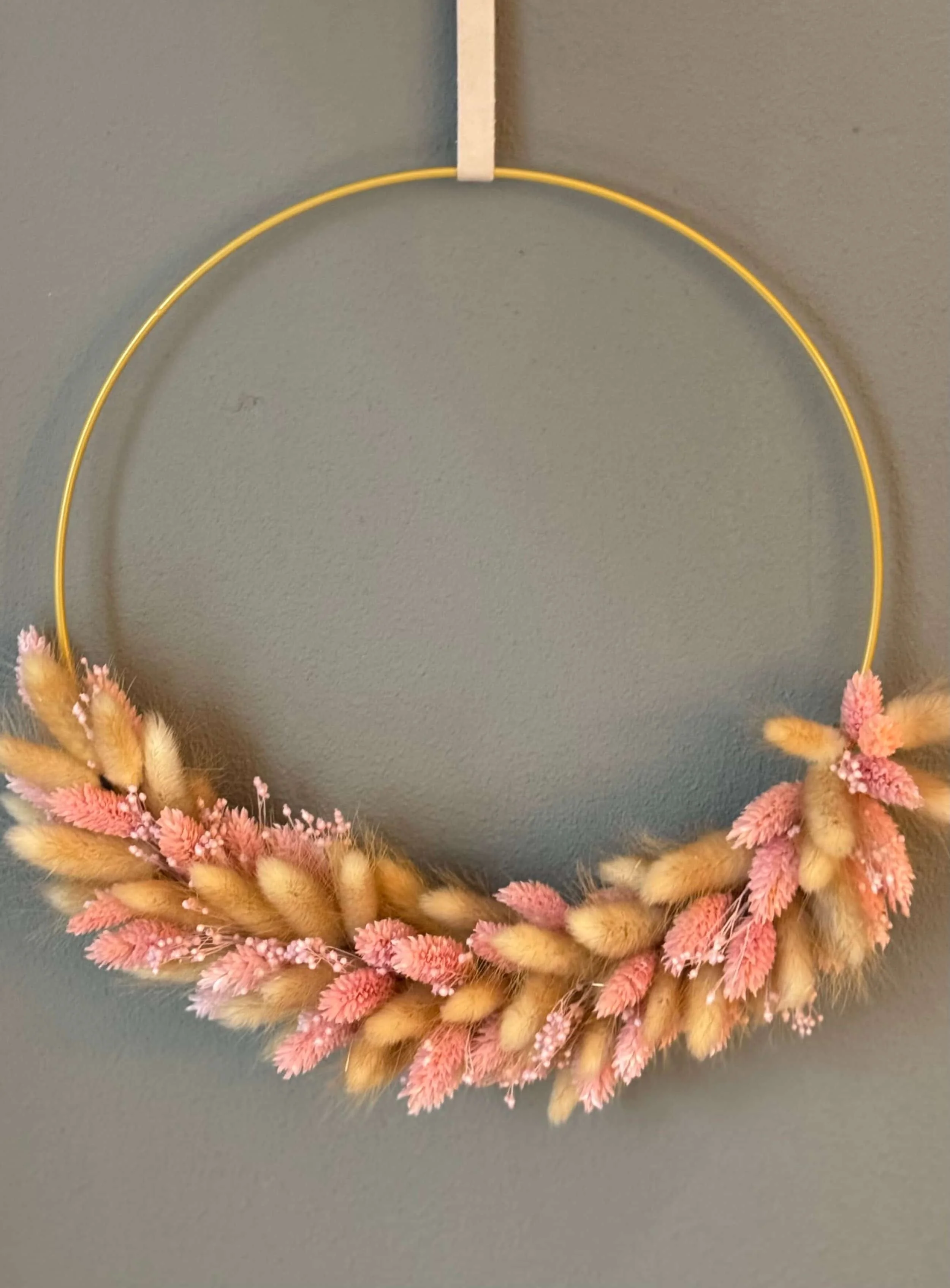 Ein kreisförmiger Blumenkranz aus getrockneten Blumen in Beige und Rosa auf einem grauen Hintergrund.