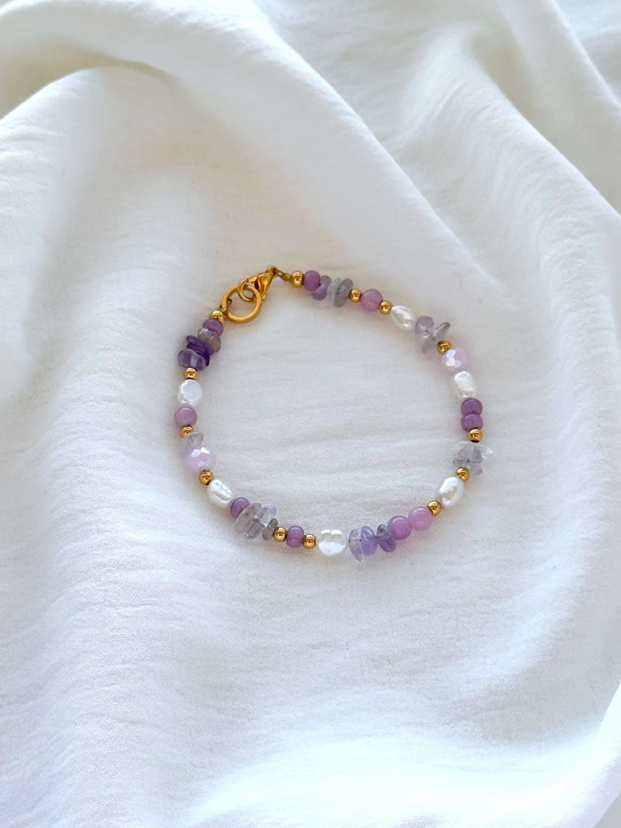 Armband aus lilafarbenen, weißen, transparenten und goldenen Perlen auf weißem Stoff