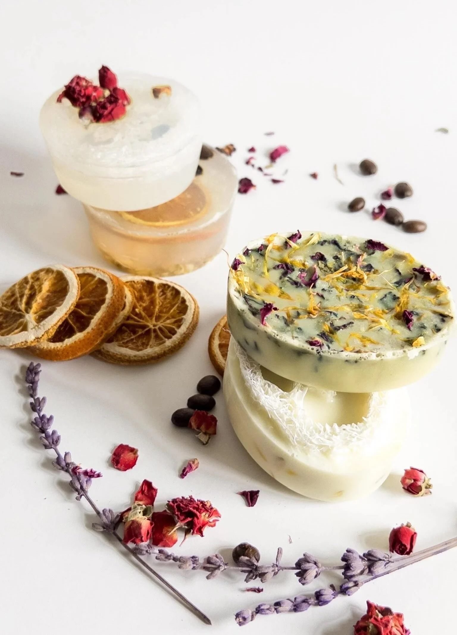 Zwei weißen Seifenstücke mit getrockneten Blumen und Kräutern, umgeben von getrockneten Orangenscheiben, Lavendel und Rosenblüten, auf weißem Hintergrund.