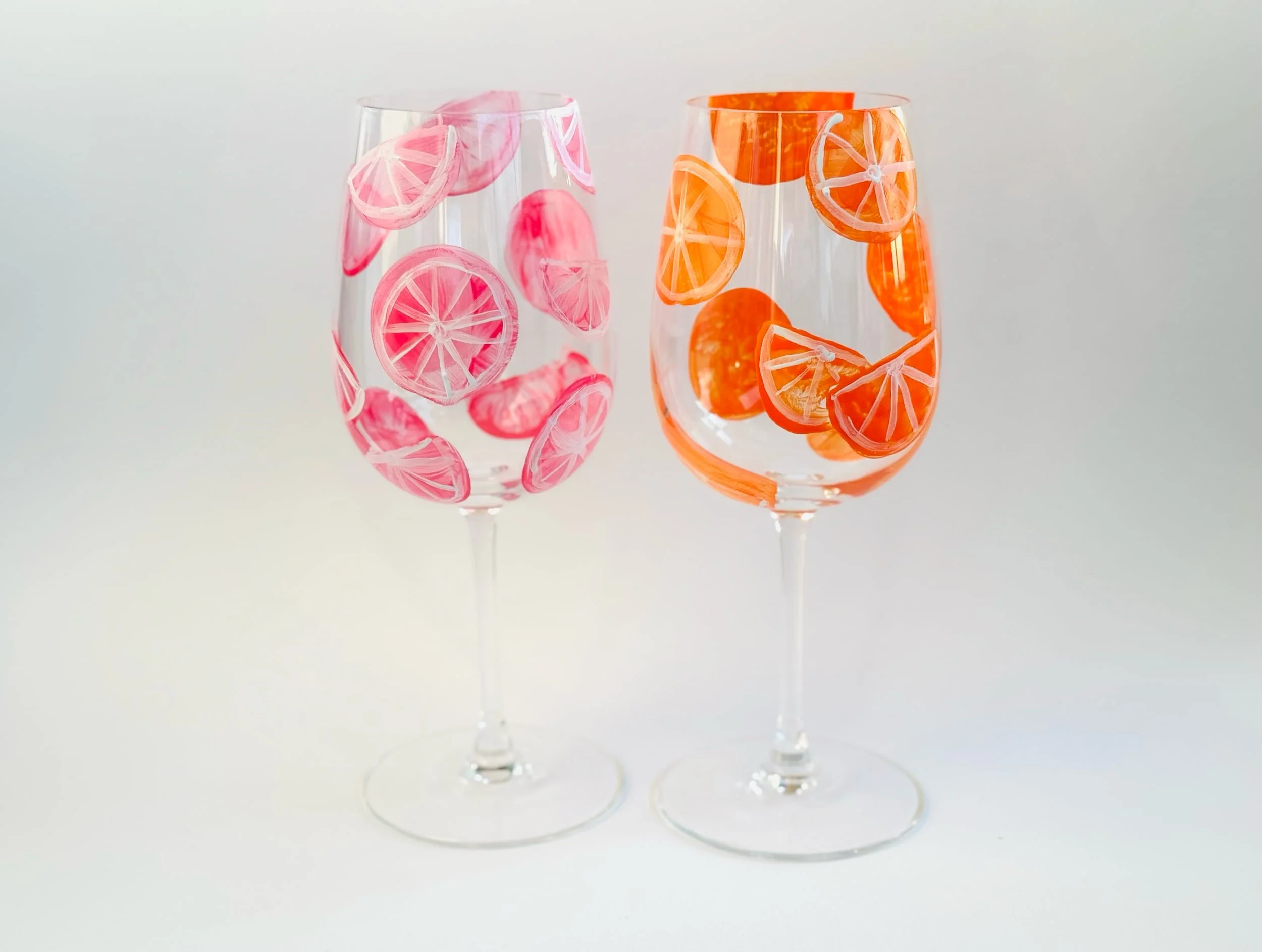 Zwei Weingläser mit Orangenscheiben-Design, eins in Pink und eins in Orange, auf weißem Hintergrund.