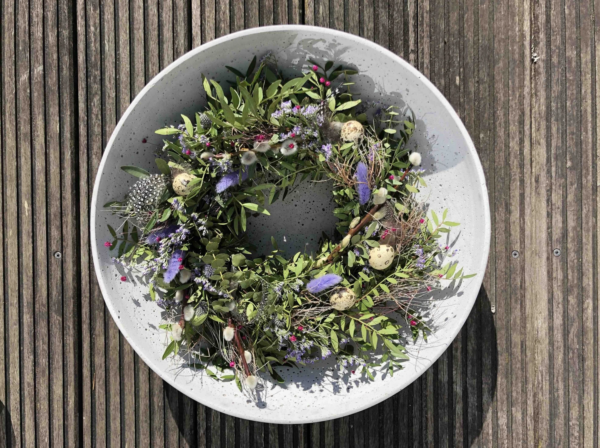Kranz aus vielen grünen Zweigen, bunten Federn, frischen und trockenen Blumen sowie kleinen Eiern, liegt in der grauen Schale auf deinem Holzuntergrund