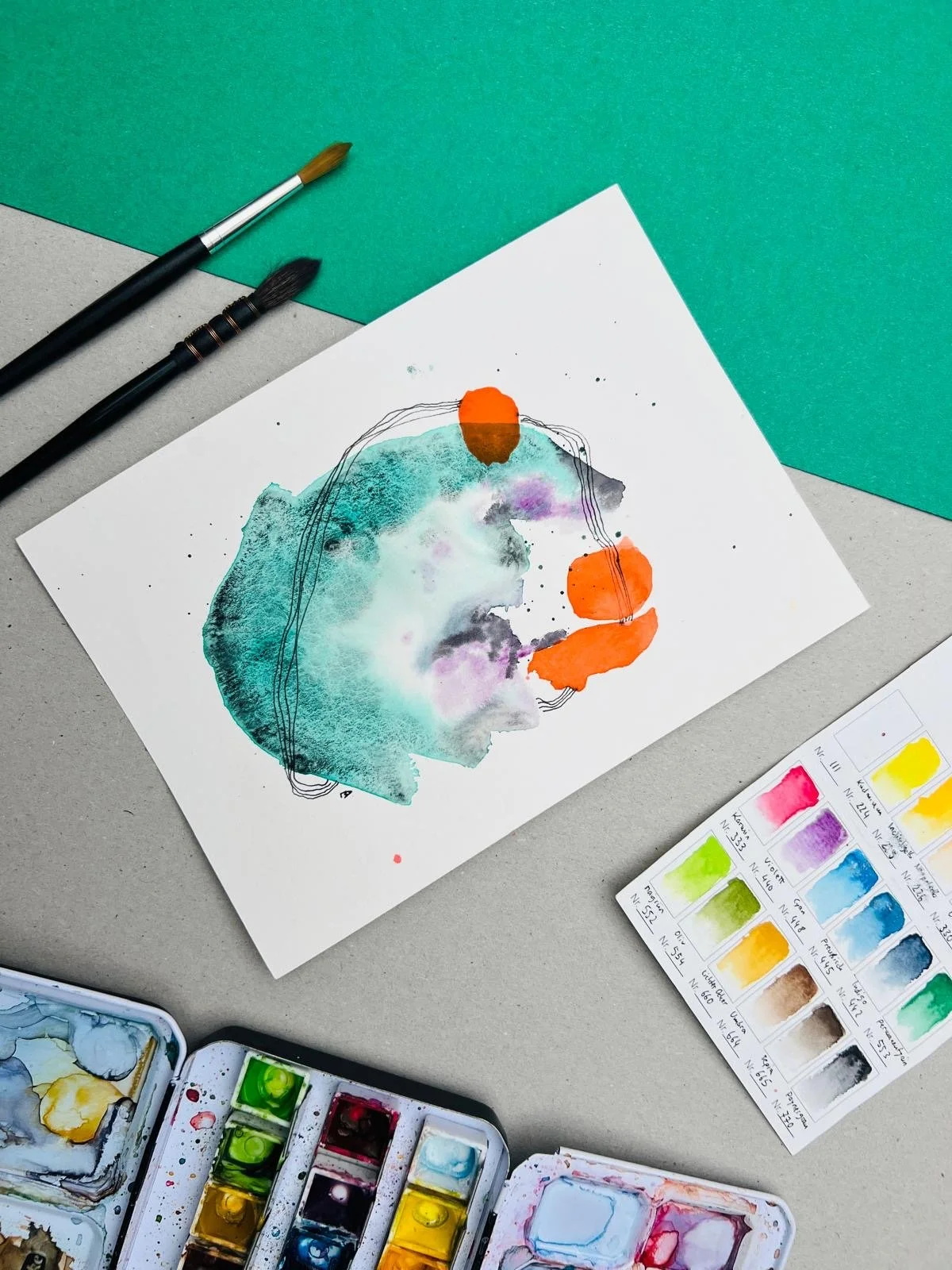 Abstraktes buntes Aquarellbild,hauptsächlihc in grün und orange sowie zwei Pinsel, zwei Aquarellkasten