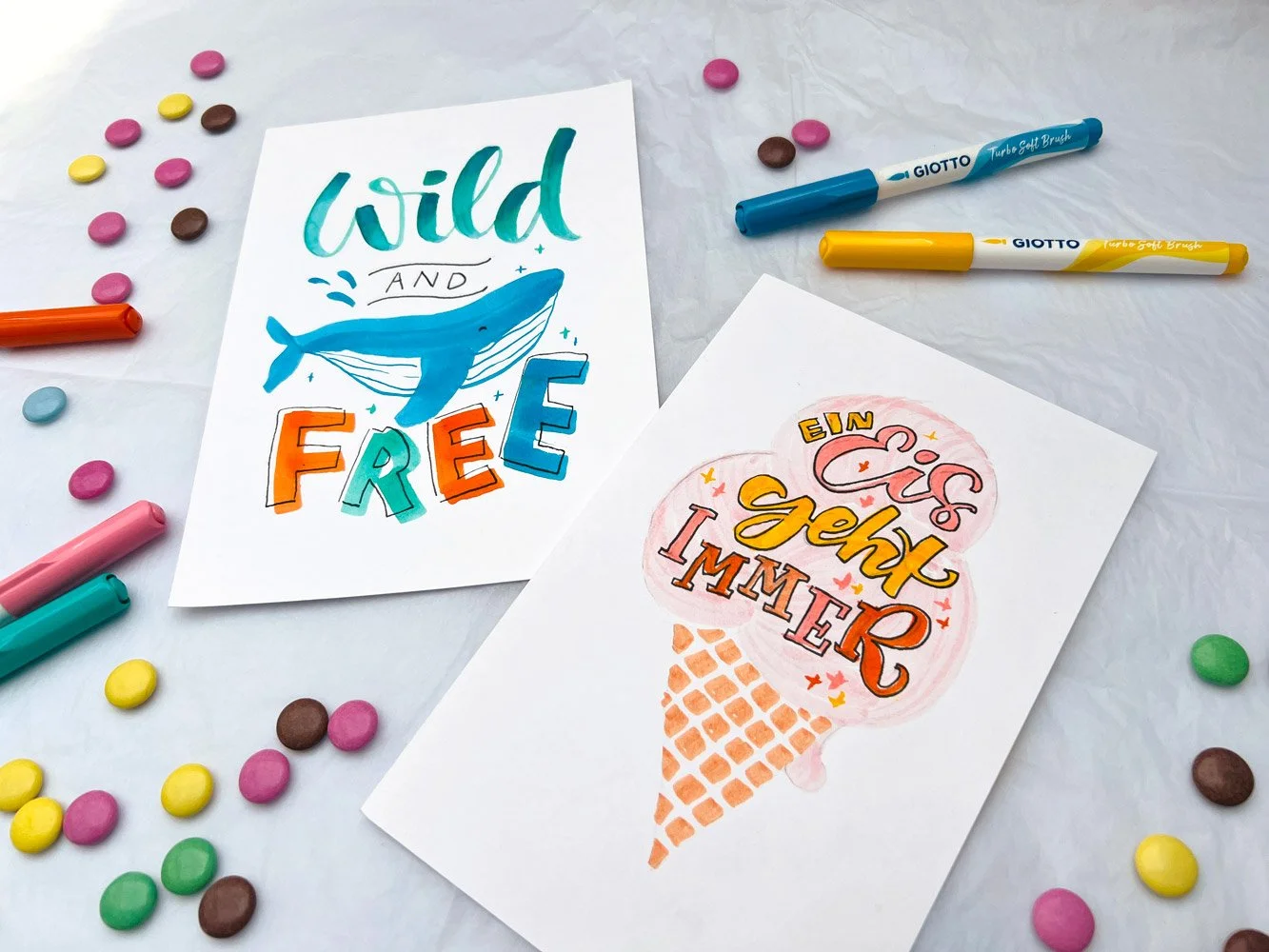 Selbstgestaltete, bunte Karten mit Lettering umgeben von Stiften und bunten Linsen