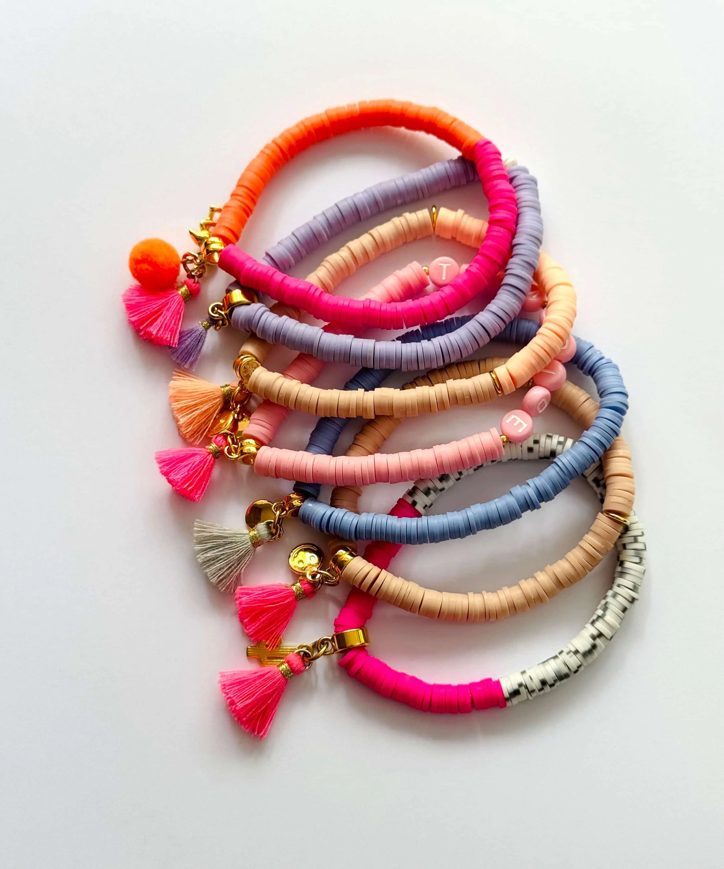 Mehrere farbige Katsuki-Armbänder mit Anhängern und Quasten, in Pastellfarben wie rosa, beige, lila, blau, und grau, auf weißem Hintergrund.