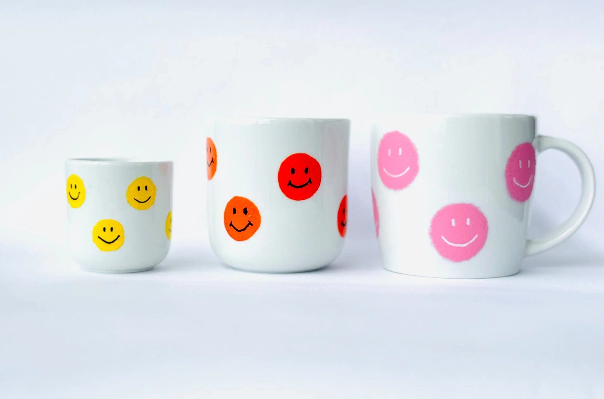 Drei selbstbemalte, weiße Tassen mit lachenden Smileys in Gelb, Orange und Pink auf weißem Hintergrund.