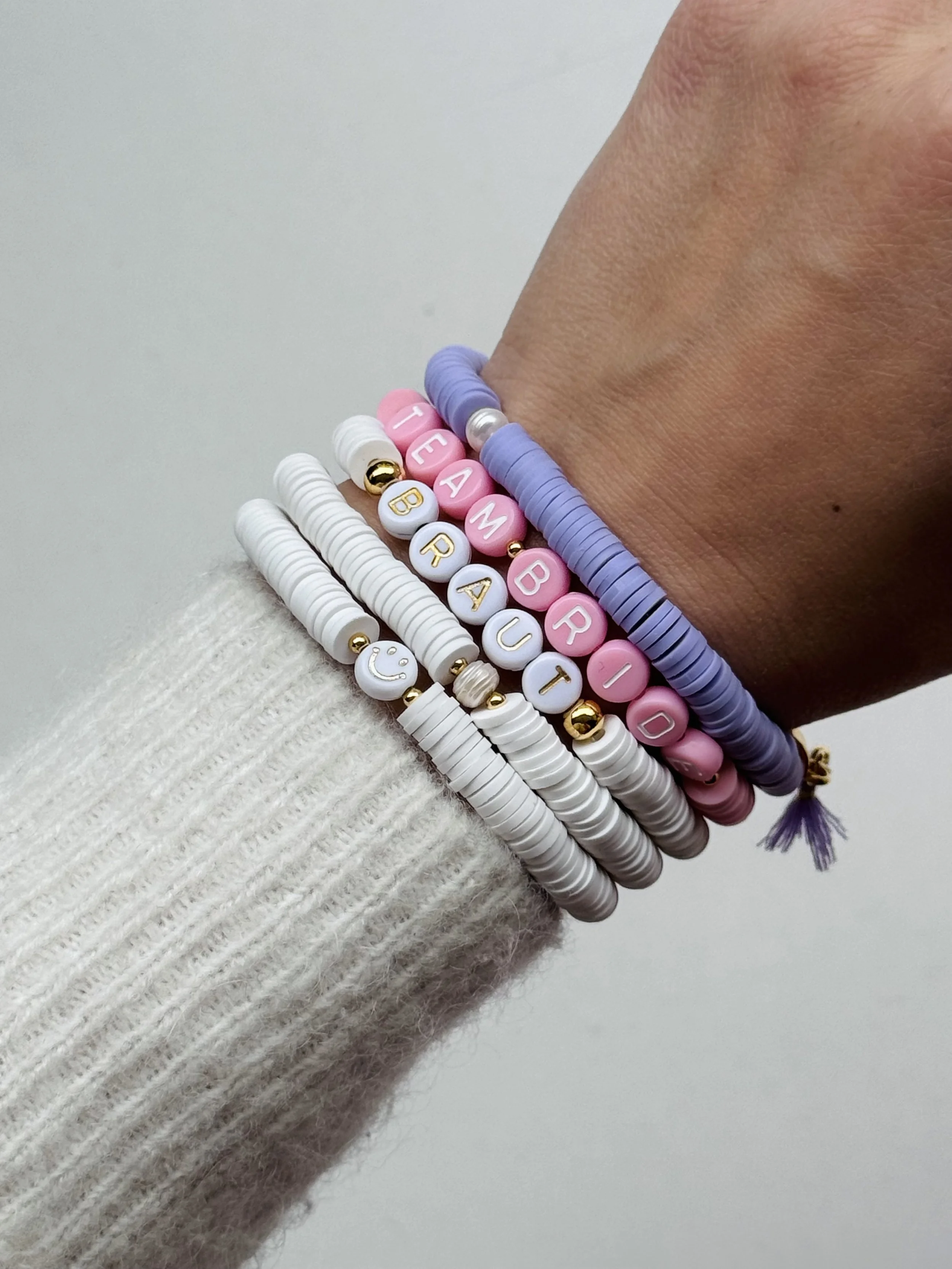 Ein Handgelenk mit mehreren bunten Armbändern, das die Worte "TEAM BABY" und einzelne Buchstaben per Perlen bildet, in Rosa, Weiß und Lila.