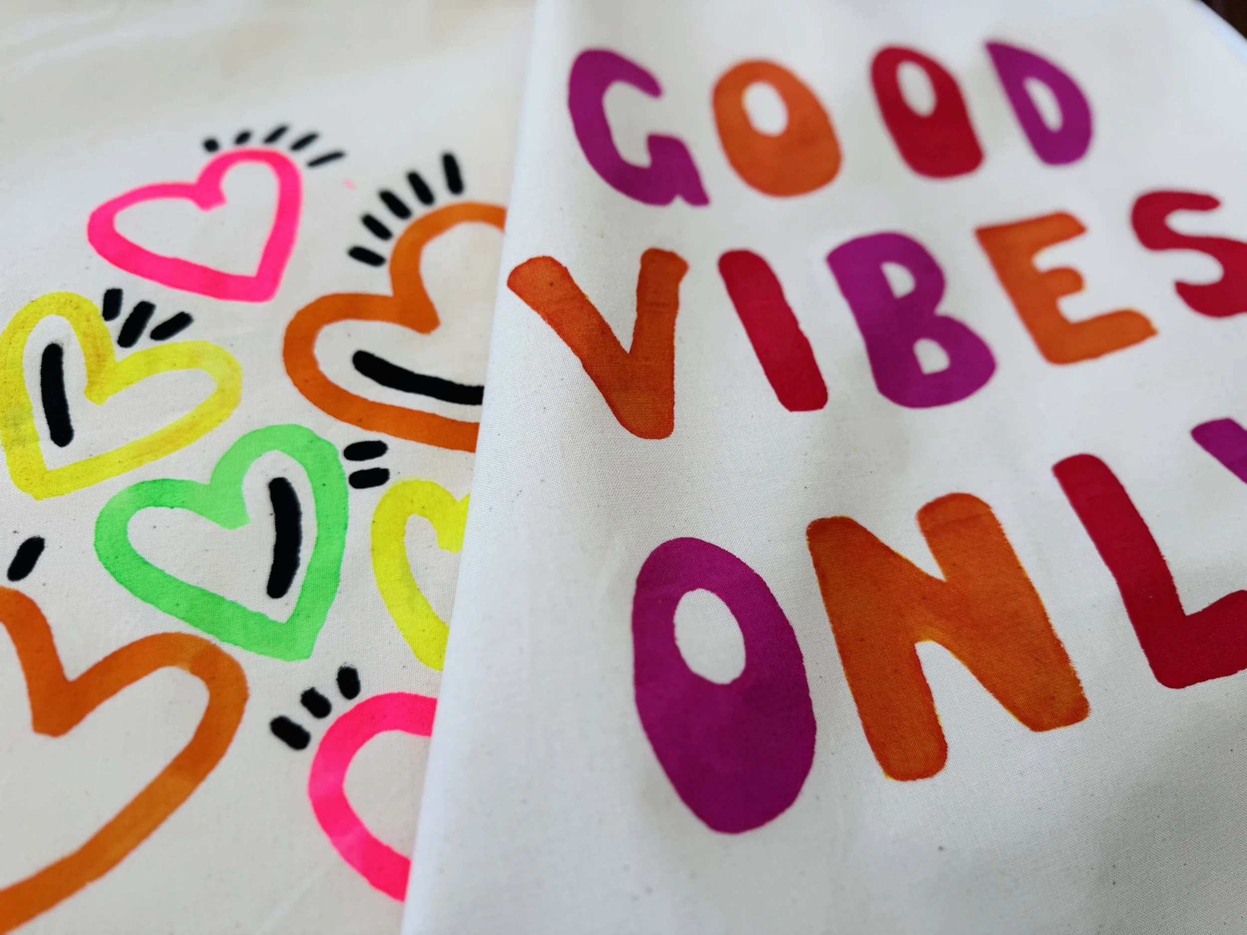 Hochwertige bunte Handzeichnungen mit Herzen und dem Text 'Good Vibes Only' auf Textilstoff, in Nahaufnahme.