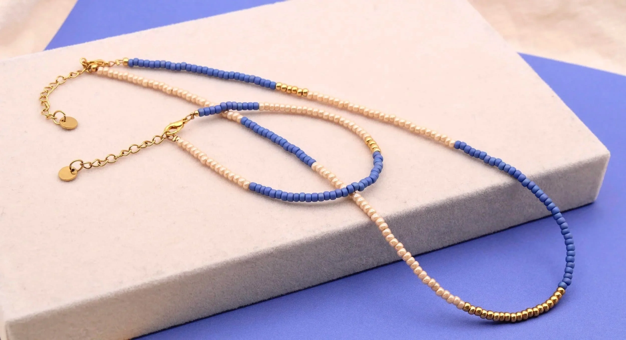 Glasperlen Armband und Kette in blauen und beigen Perlen auf beigem Brett und blau-lila Untergrund