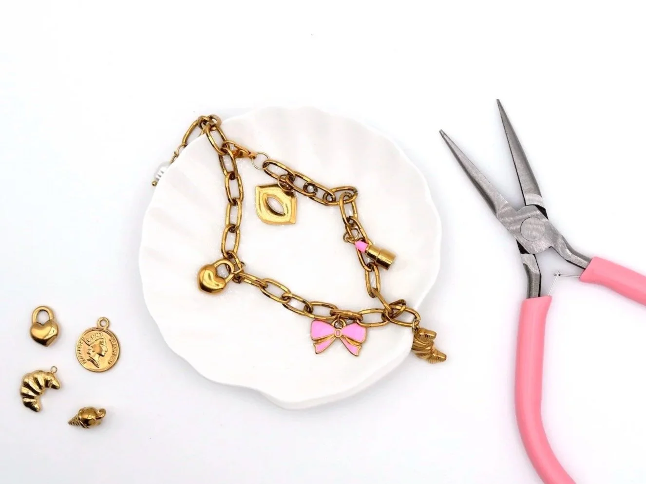 goldene Gleiderkette mit Anhänger Herz in gold, Scheleife in rosa, Mund in gold und Lippenstift in gold und rosa, liegend auf einer weißen Schale in Form einer Muschel. Daneben eine Zange in rosa 