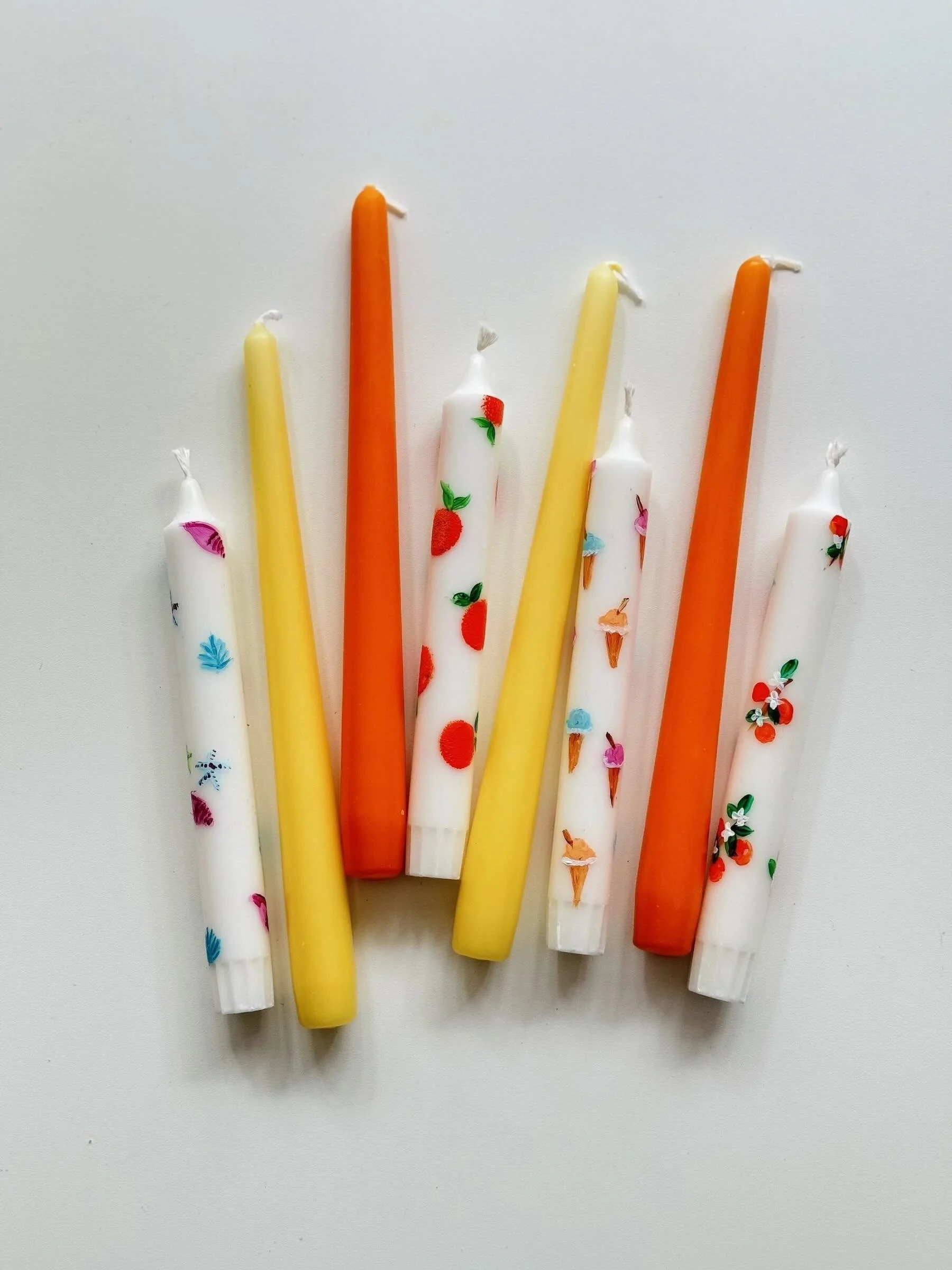 Bunte Spitzkerzen und weiße Stabkerzen bemalt mit Motiven wie Orangen, Eis in der Waffel, Mandarinen, Muscheln
