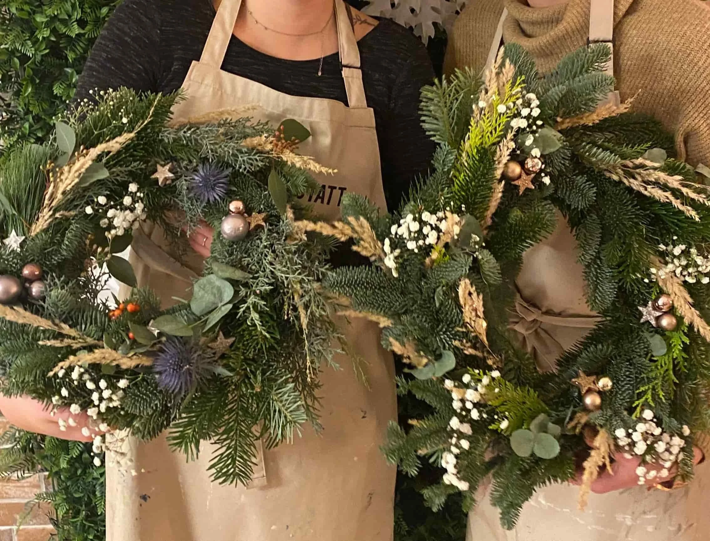 zwei Personen in beigenen Schürzen halten vor sich WEihnachstkränze bestens aus Tanne, Kiefer, Eukalyptus, weißem Schleierkraut und Kugeln