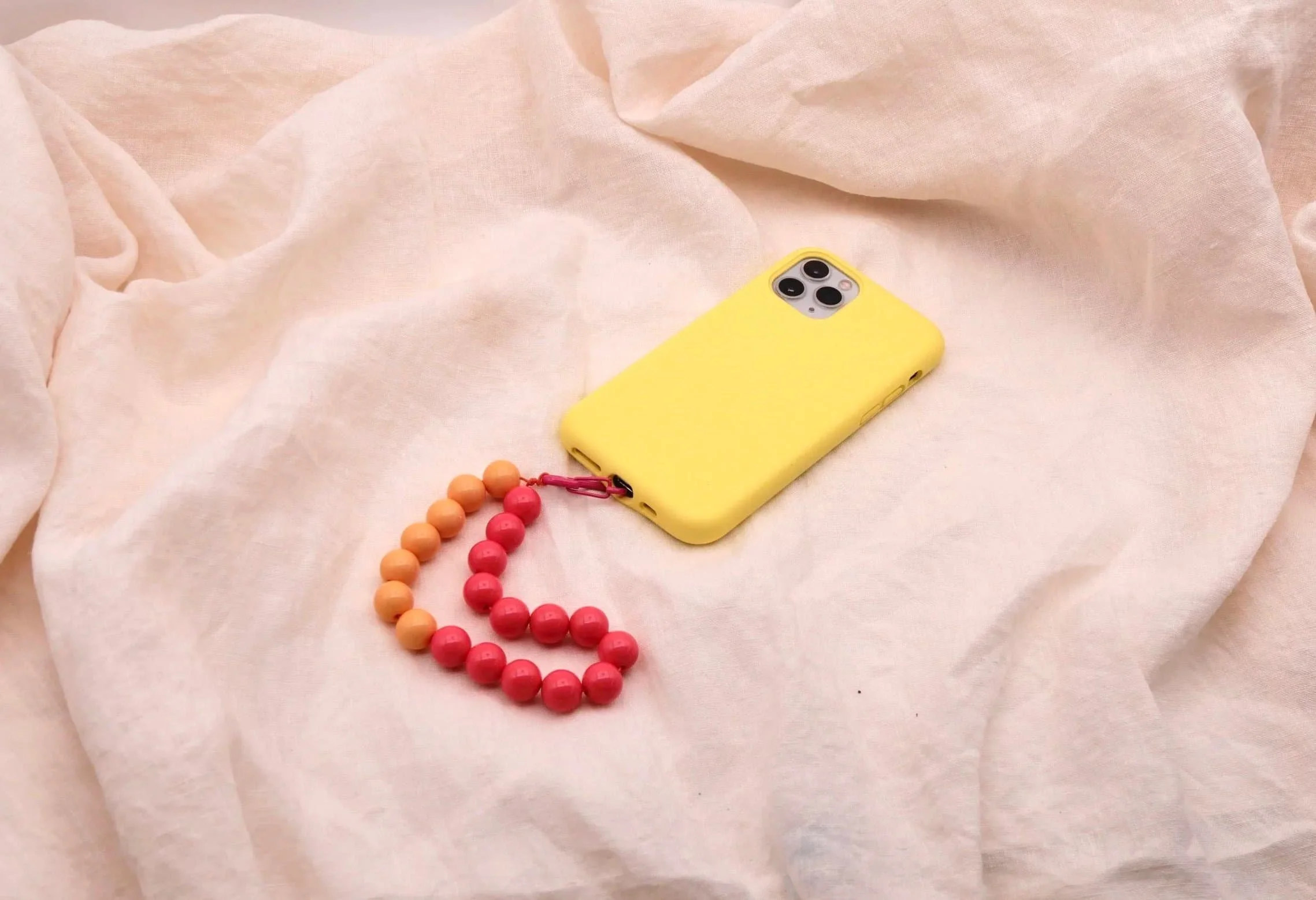 Ein gelbes Smartphone mit einer Handyhülle liegt auf einem hellrosa, weichen Stoff. An der Hülle ist eine Kette mit roten und orangenen Holzperlen befestigt.