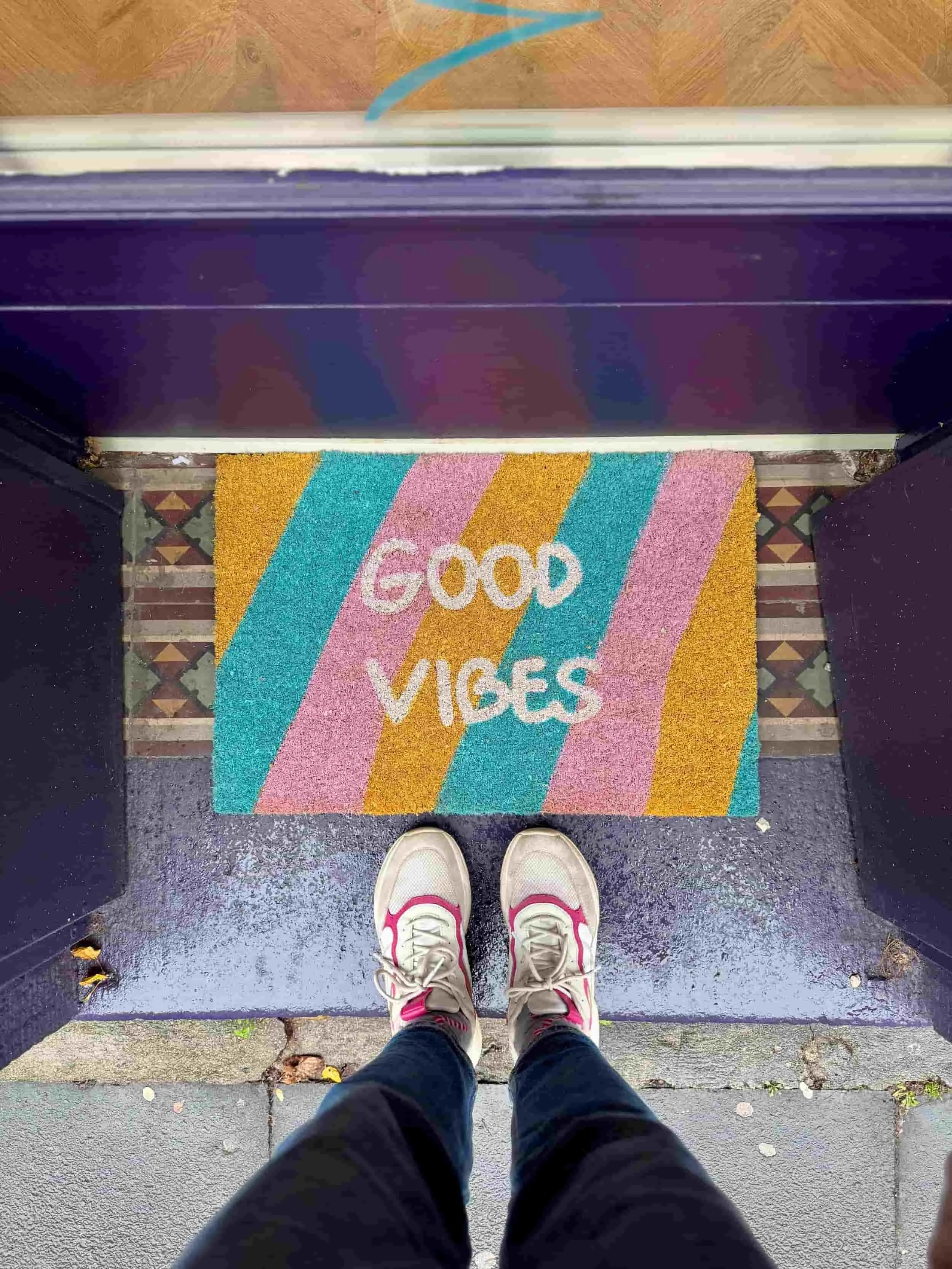 Eingangsbereich mit einer bunten Fußmatte in türkis, gelb und rosa mit den Worten Good Vibes in weiß sowie einem paar Füße mit beige-pinken Sneakern