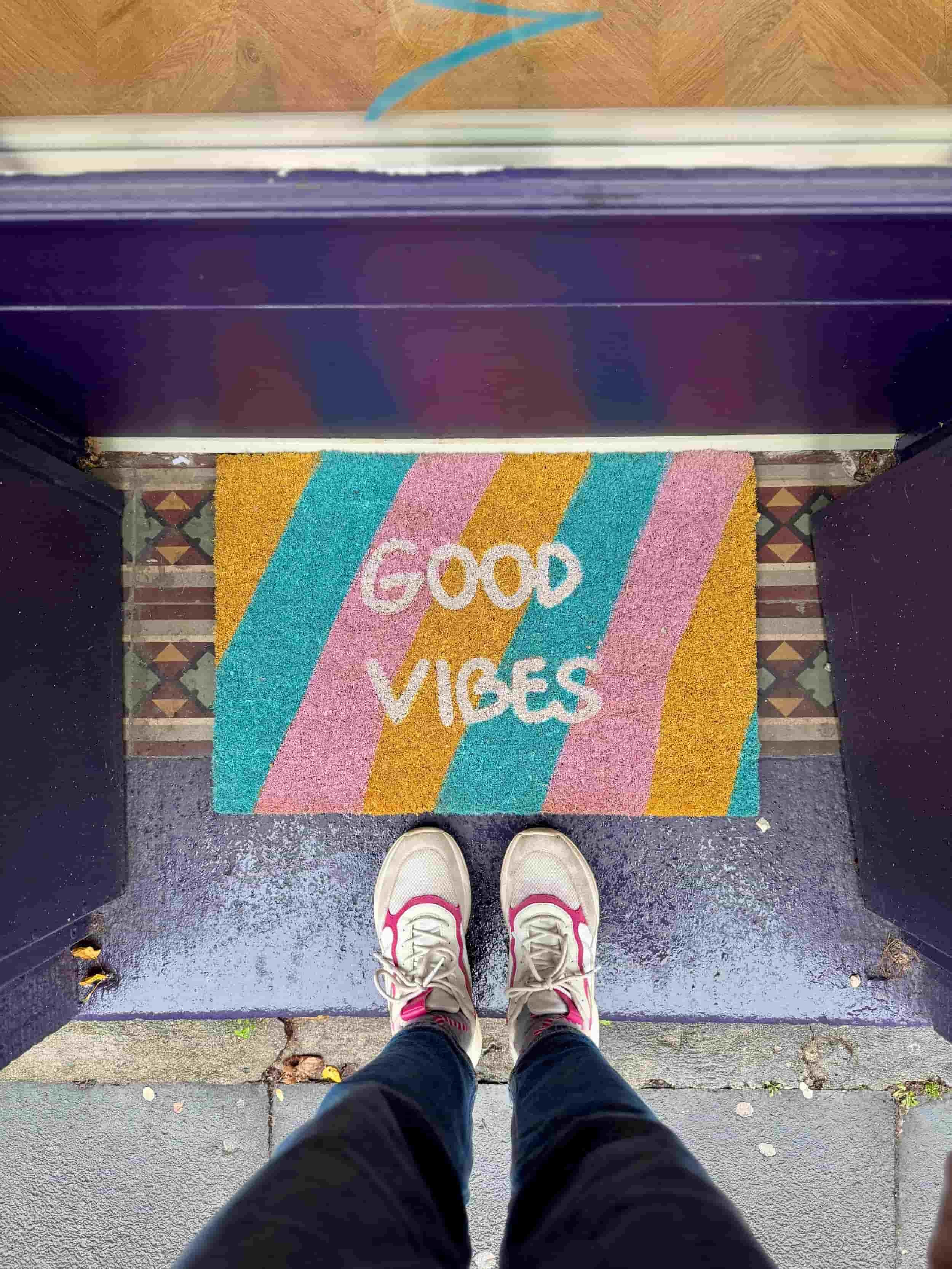 Eingangsbereich mit einer bunten Fußmatte in türkis, gelb und rosa mit den Worten Good Vibes in weiß sowie einem paar Füße mit beige-pinken Sneakern