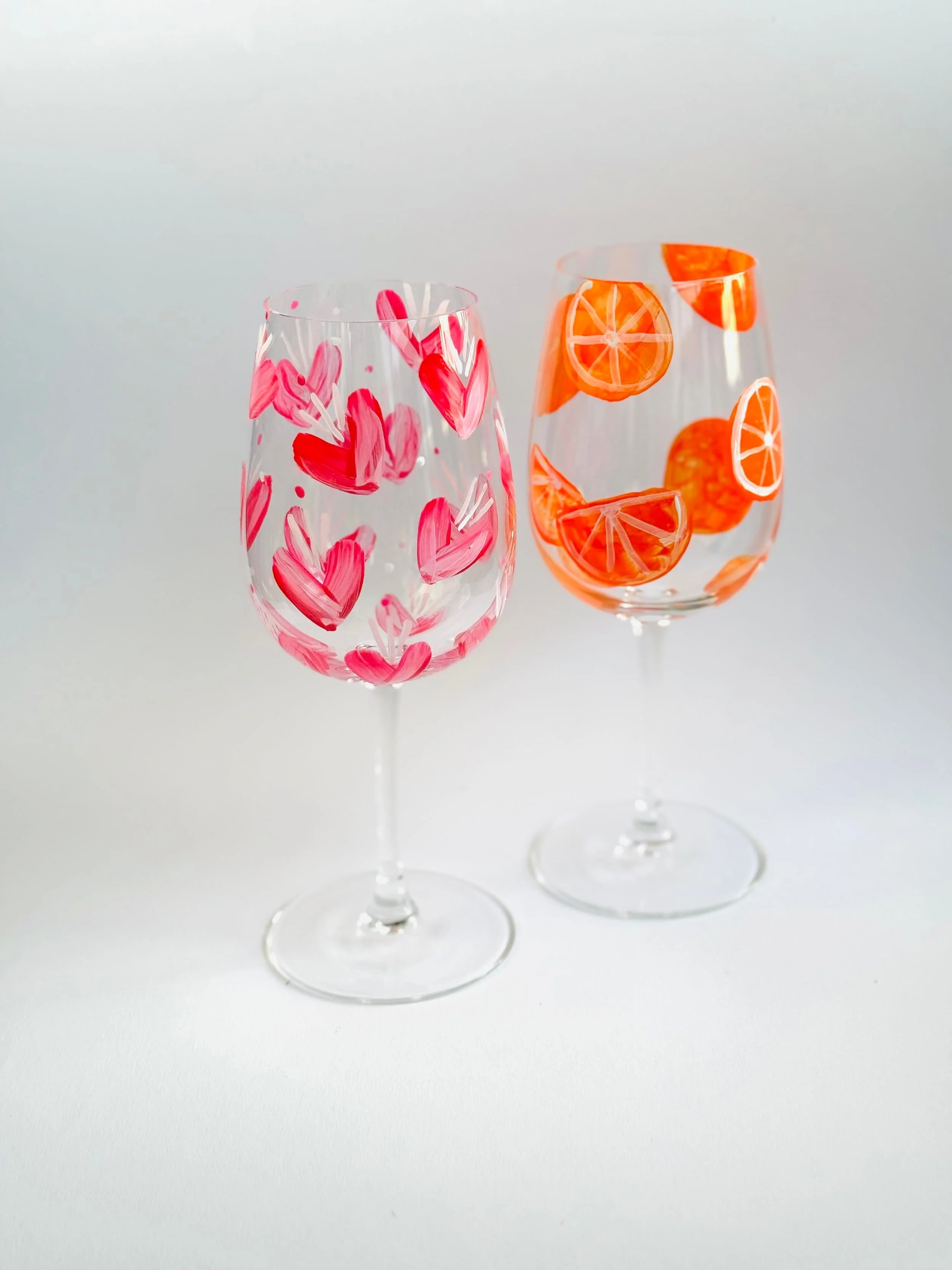 Zwei Weingläser mit roten Herzen und Orangenscheiben, handbemalt, auf weißem Hintergrund.