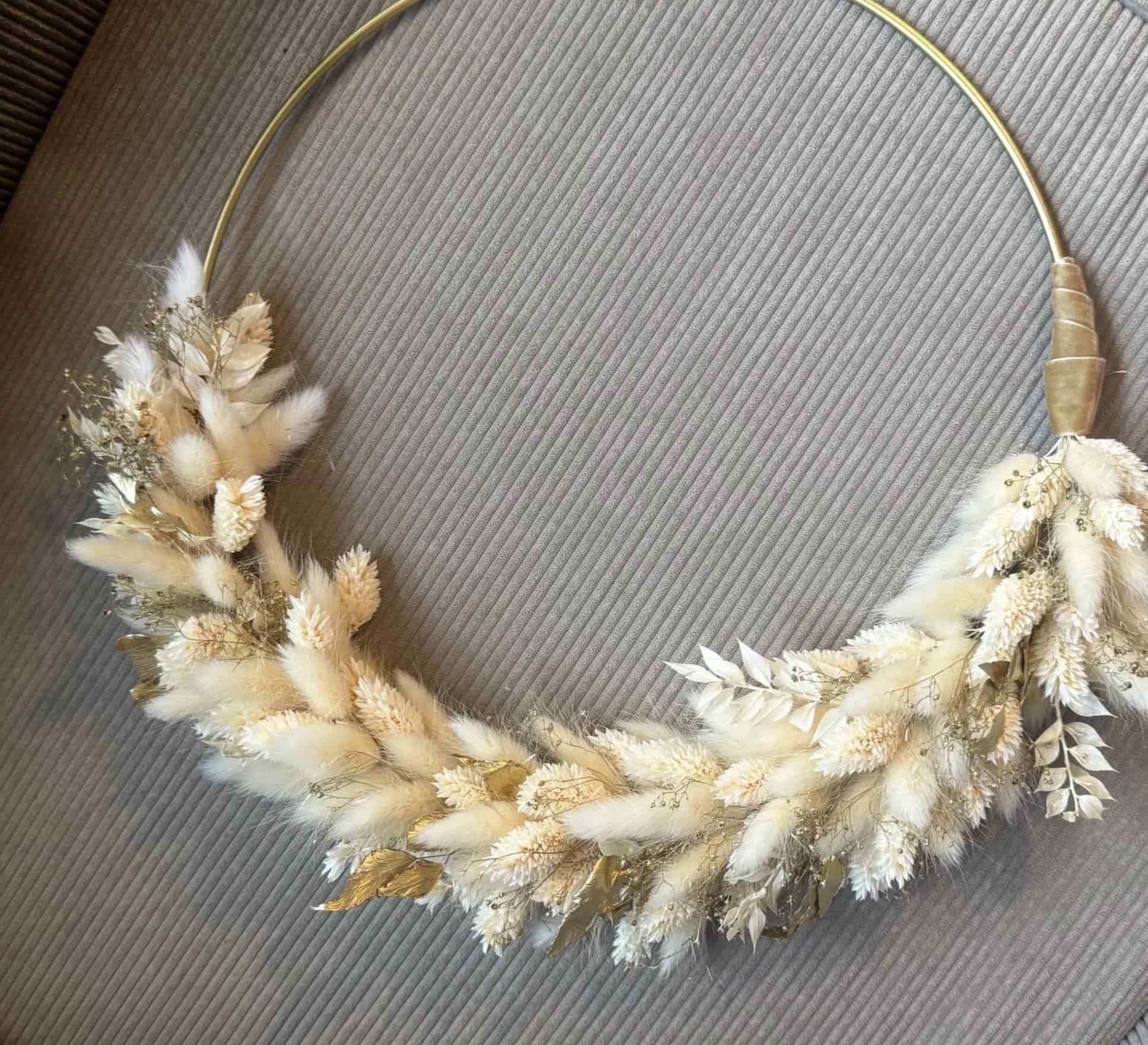 Blumenkranz mit weißen und goldenen Trockenblumen und Blättern, an einem runden goldenen Draht angebracht.