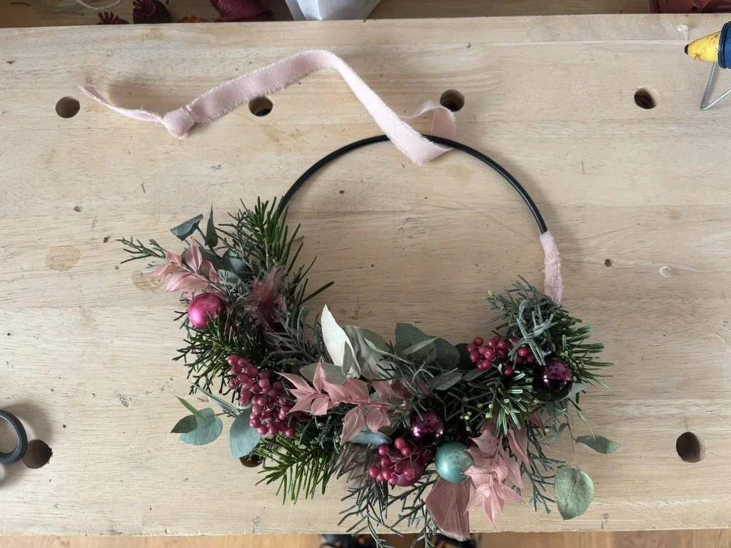 Weihnachtslicher Flower Hoop liegend auf einem Holztisch, bestehend aus Tanne, rote Skimmia, rosa Ruskus, Eukalyptus, kleine grüne und pinke Weihnachtskugeln, rosa Band