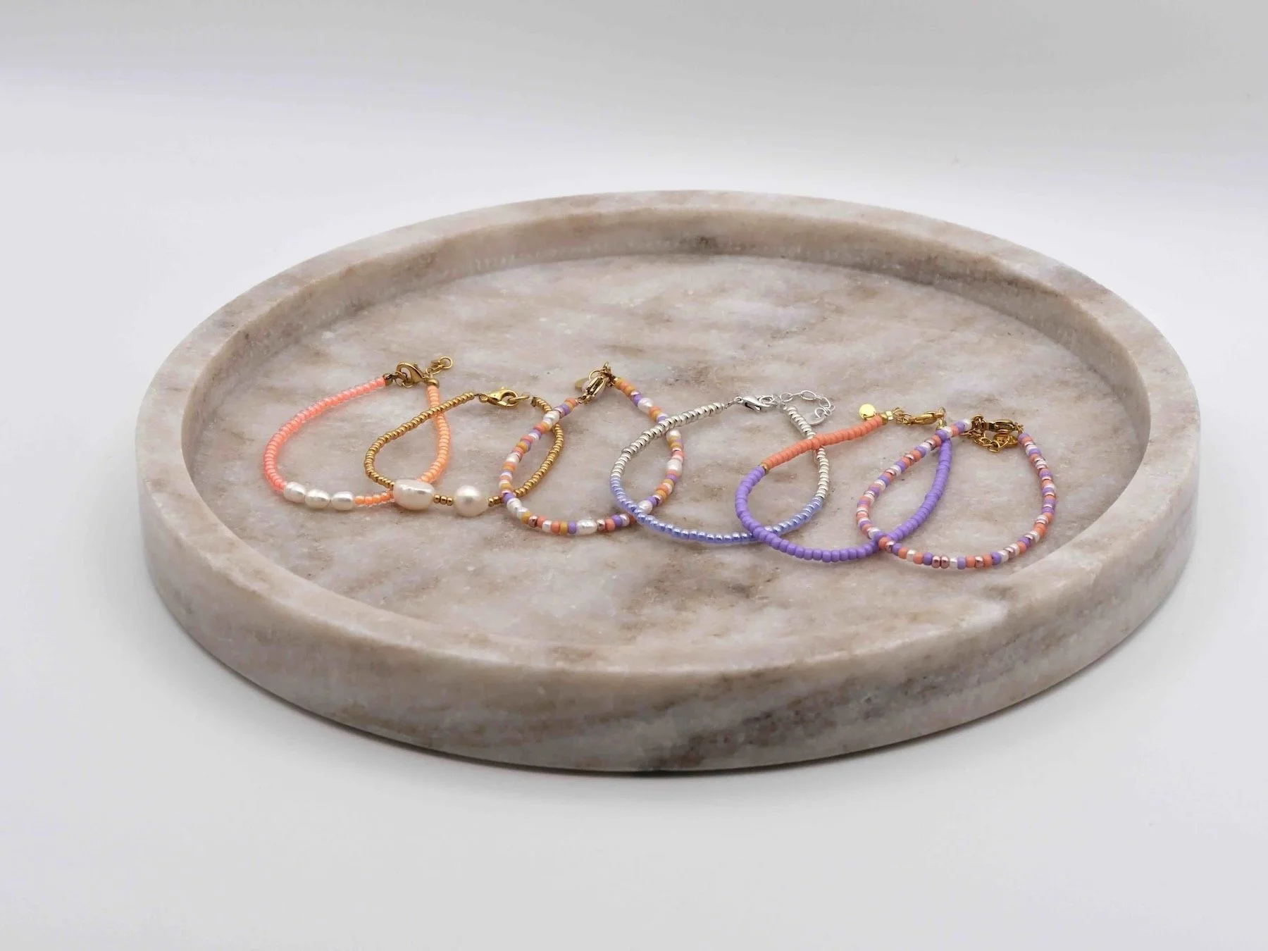 Mehrere bunte Armbänder aus Glasperlen in rosa, lila, weiß, blau, silber, gold, teilweise mit Süßwasserpelen. Armbänder liegen auf einem runden Tablett aus Marmor