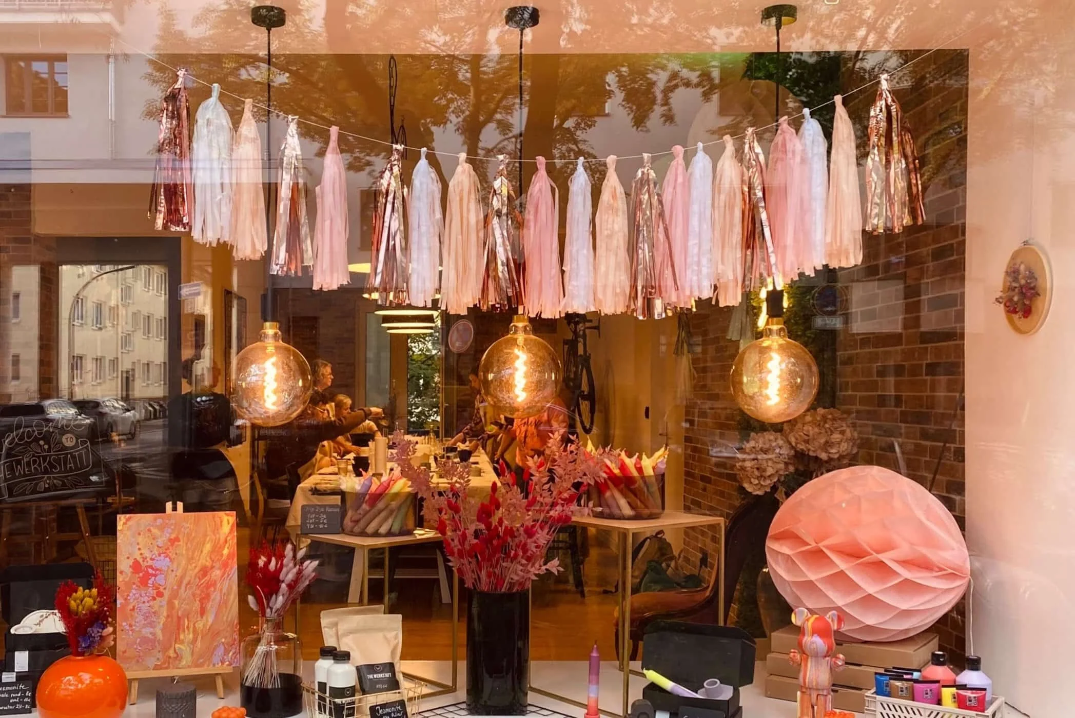 Der Blick in ein Geschäft oder einen Raum mit verschiedenen Dekorationsartikeln, darunter bunte Wände, Lampen, Blumen und Kunstwerke, die durch einen großen Fensterbereich sichtbar sind.
