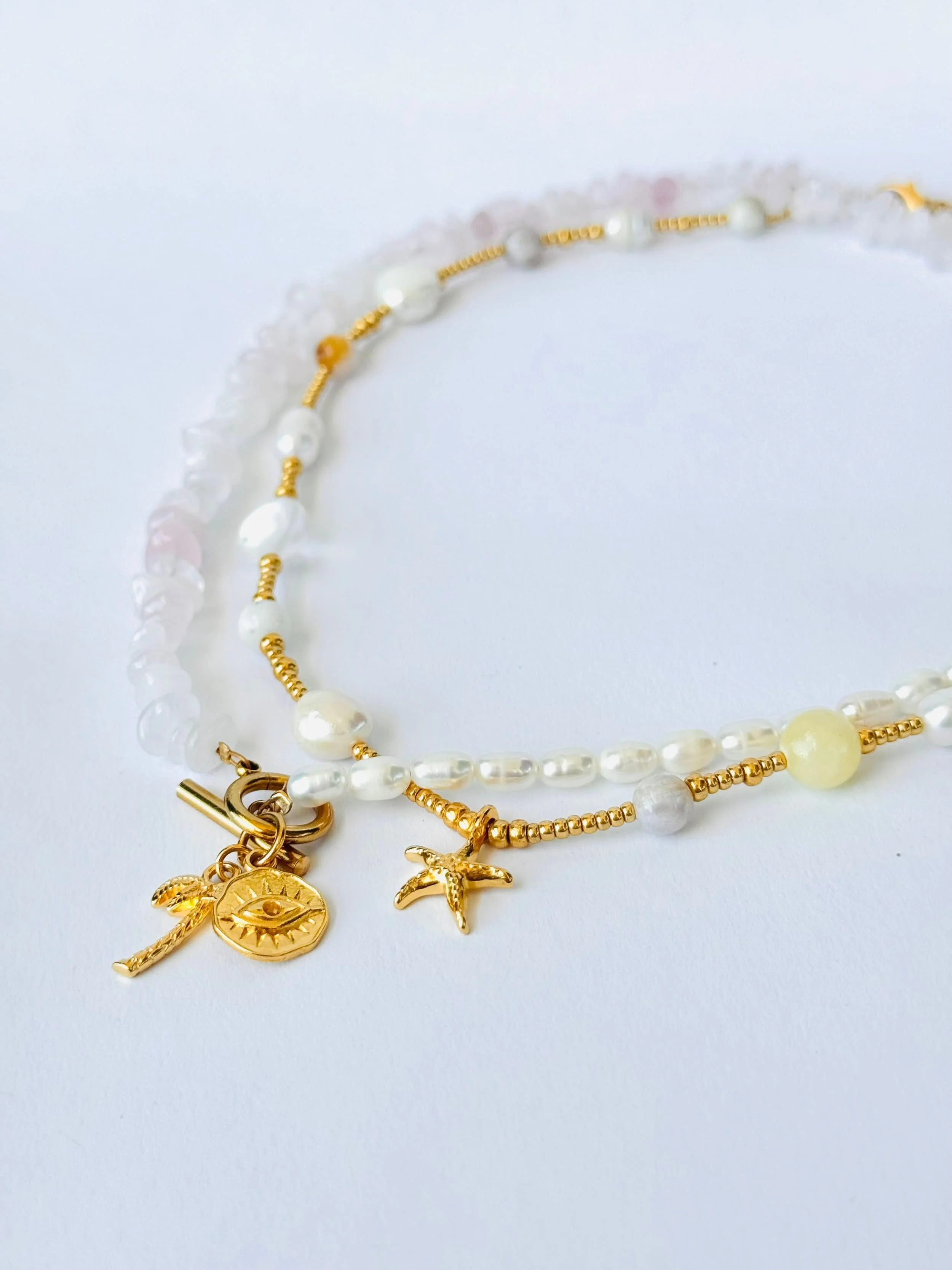 Eine Perlenkette aus Süßwasserperlen mit goldenen Anhängern, darunter ein Auge, eine Palme und eine Sonne, auf weißem Hintergrund.